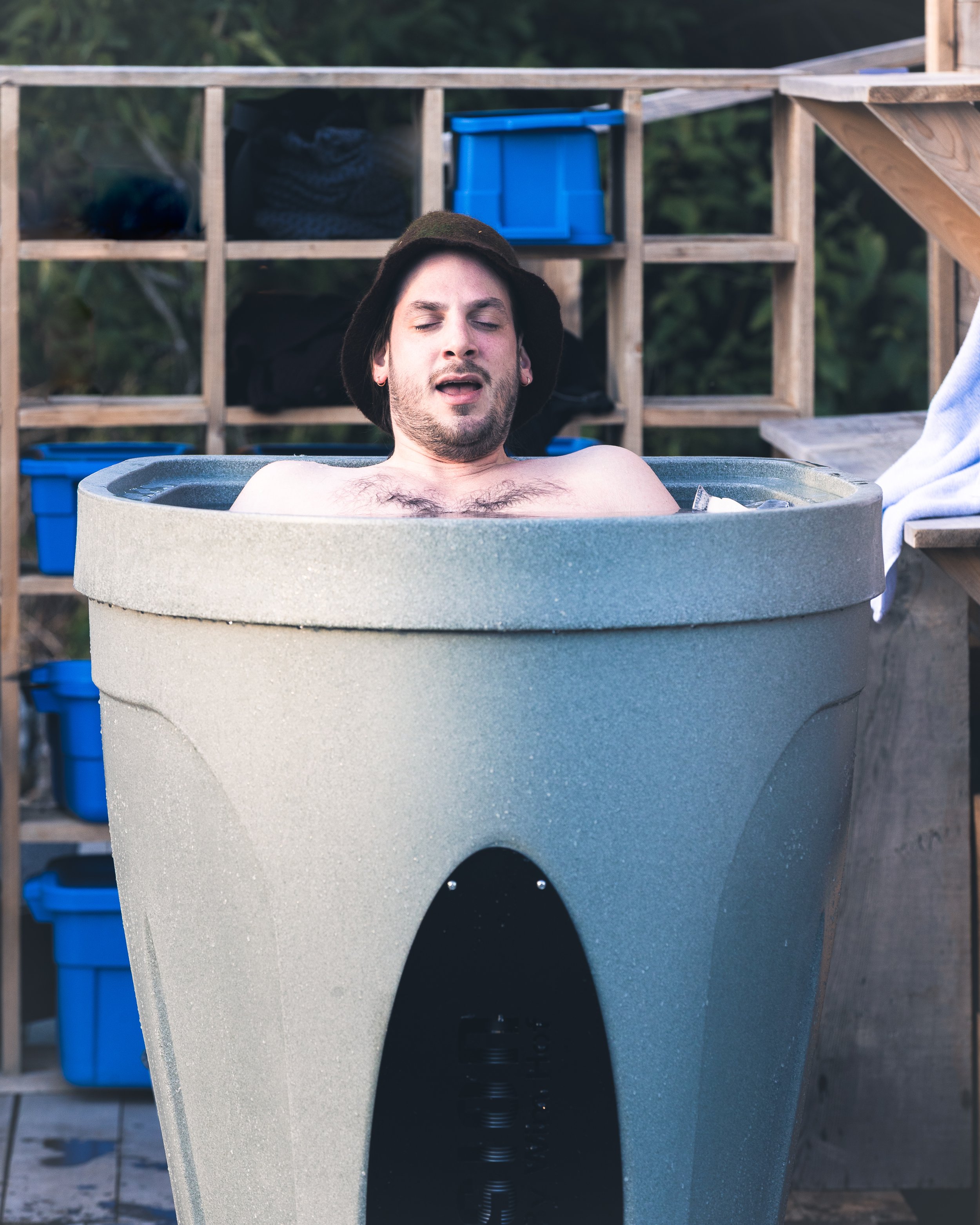 A person with dark hair, wearing earrings, relaxing in an outdoor hot tub with their eyes closed. The hot tub is located on a deck with a wooden lattice structure in the background, which has storage boxes and bags. The person is wearing a hoodie, an