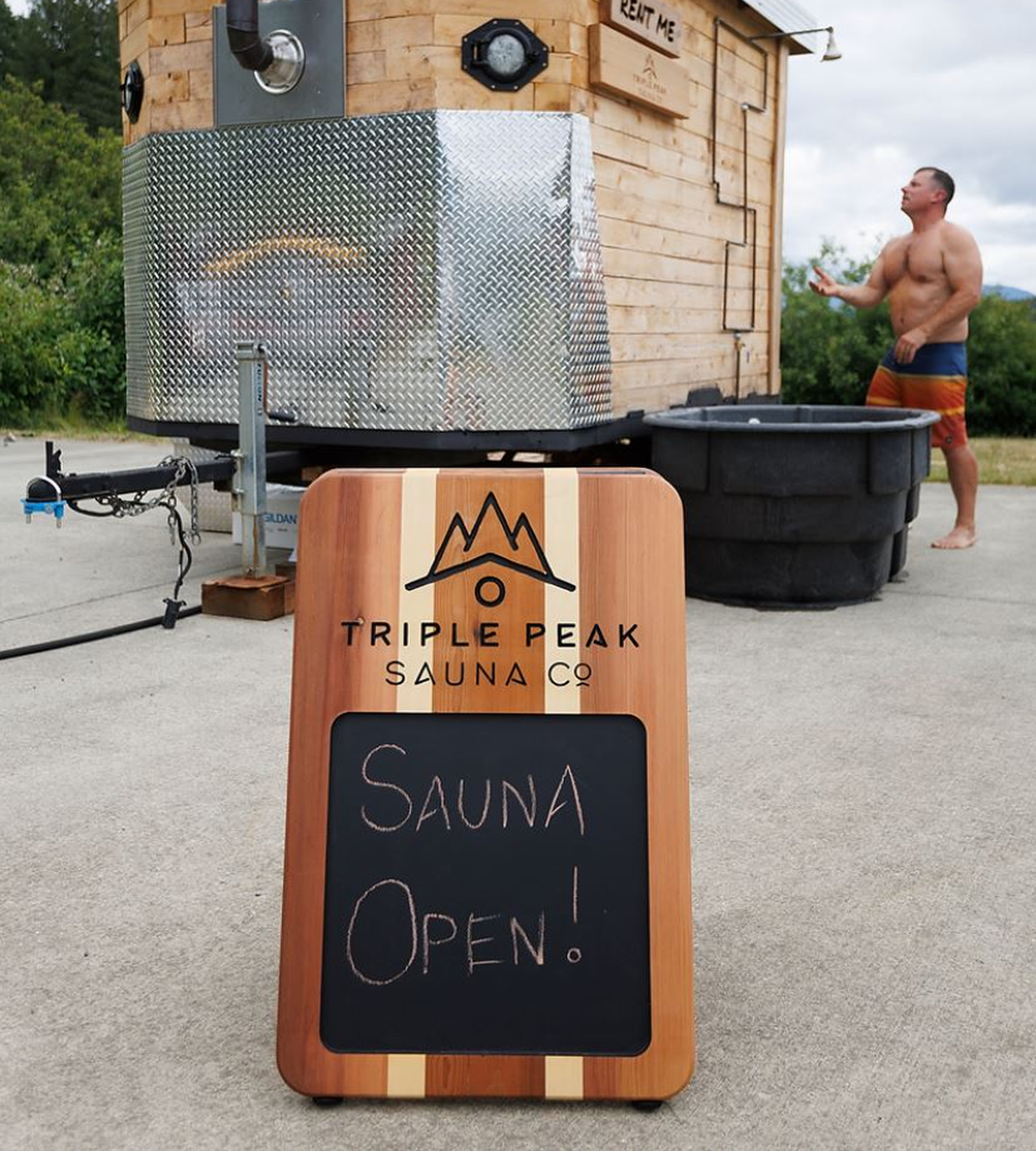 A wooden sign that reads 'Triple Peak Sauna Co. Open,' placed in front of a black container, along with a trailer and a large metal sauna on wheels. A shirtless man in colorful shorts stands nearby.