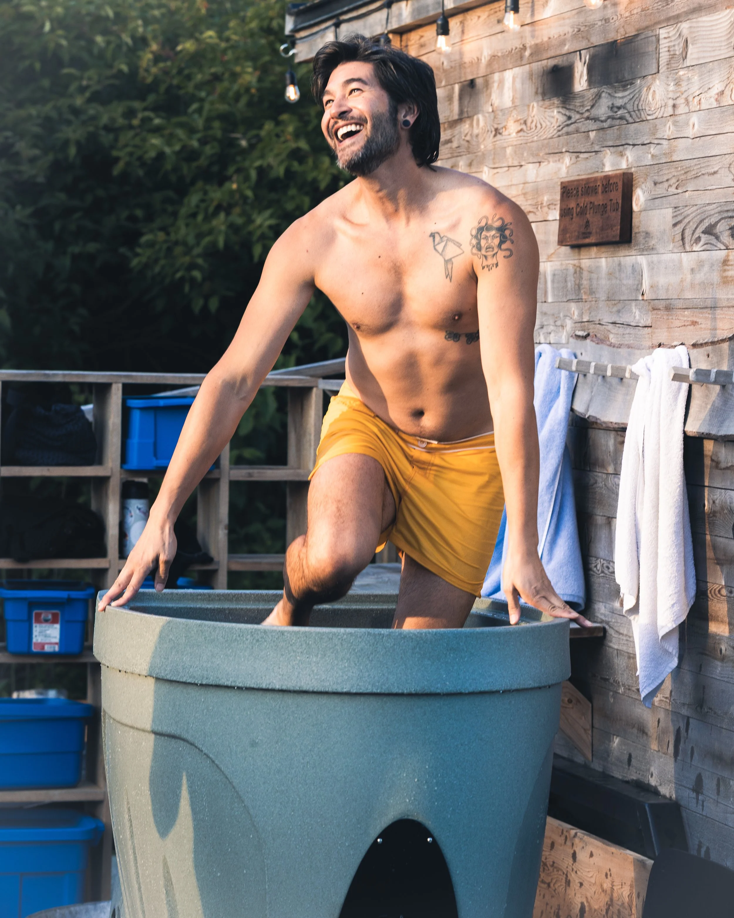 A man with dark hair and a beard, wearing yellow shorts, is smiling while sitting in a hot tub on a deck near a wooden wall. There are towels hanging on a wooden rail and various containers on shelves nearby. The background features greenery and hang