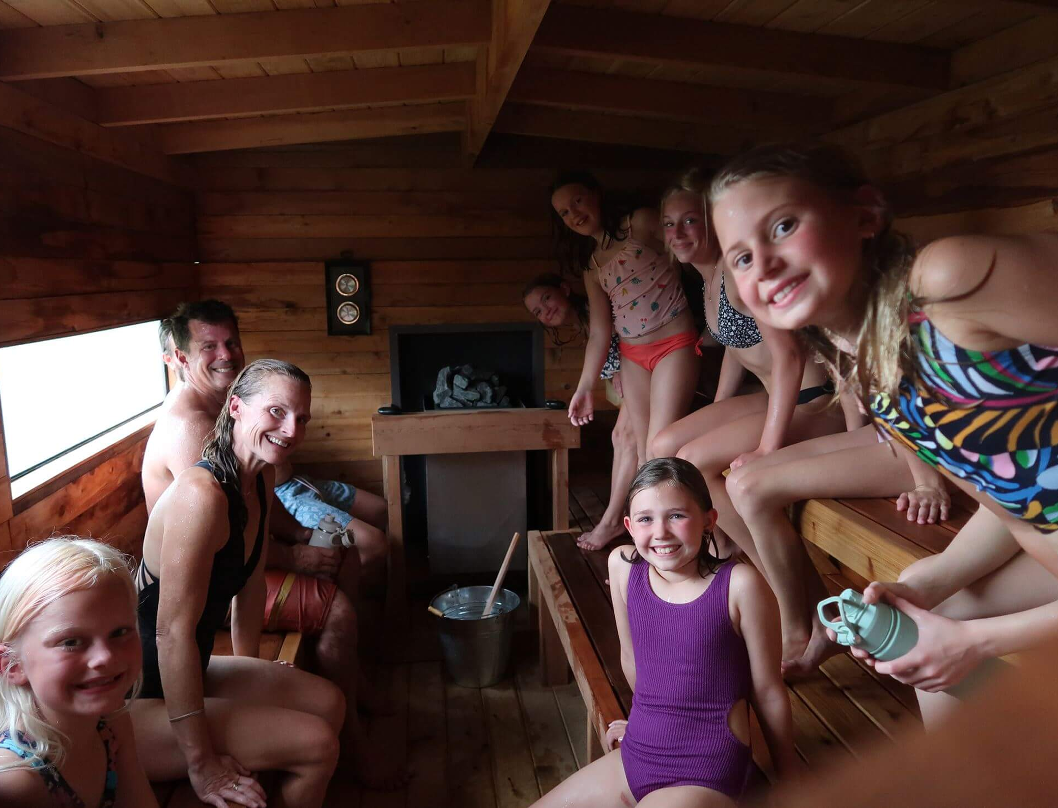 A group of children and adults inside a wooden sauna, some are smiling and sitting on the benches, with a bucket and water ladle in the center.