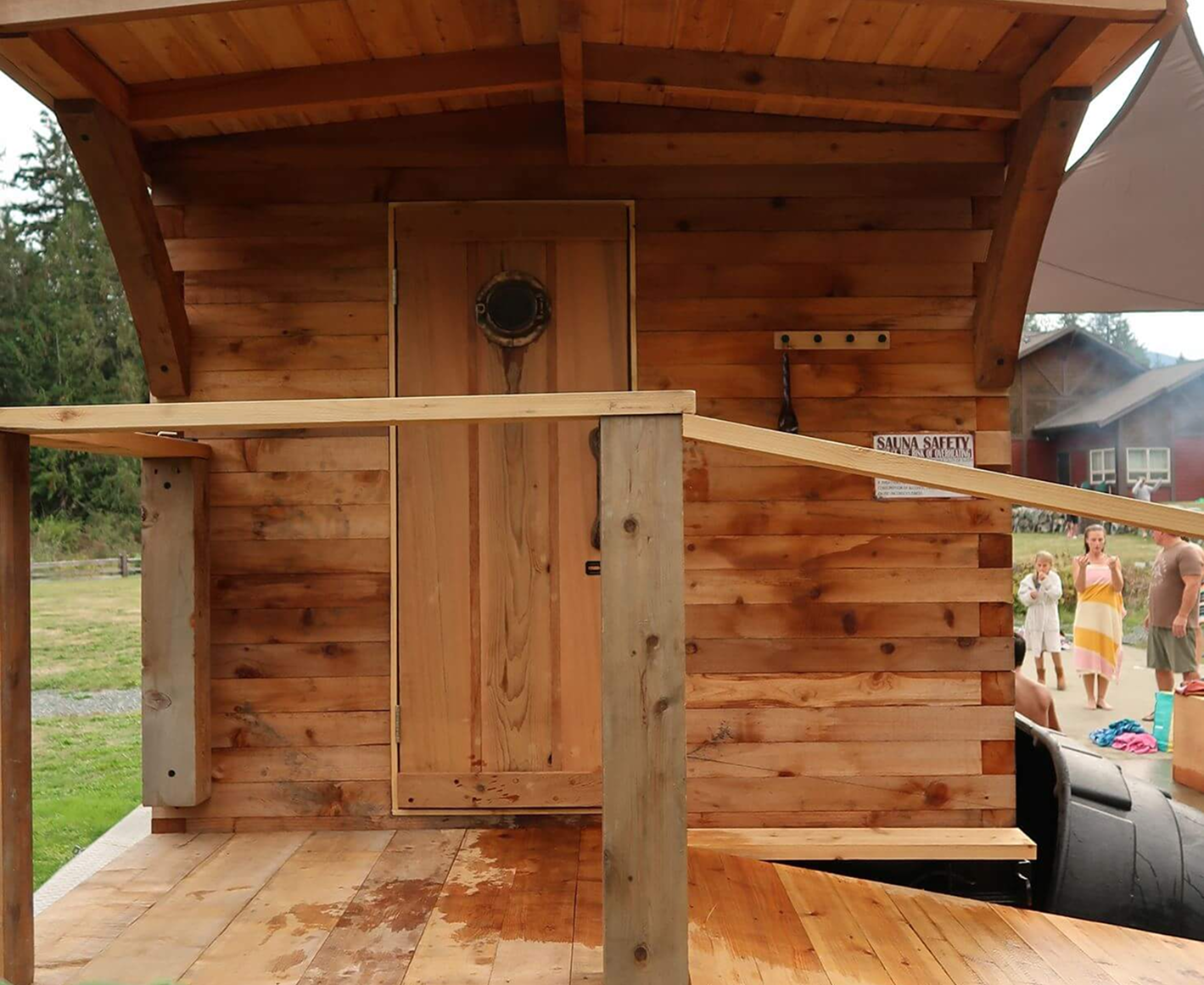 Wooden sauna cabin with a small door, partially built indoor or outdoor deck, with safety signs in the background and people standing nearby.
