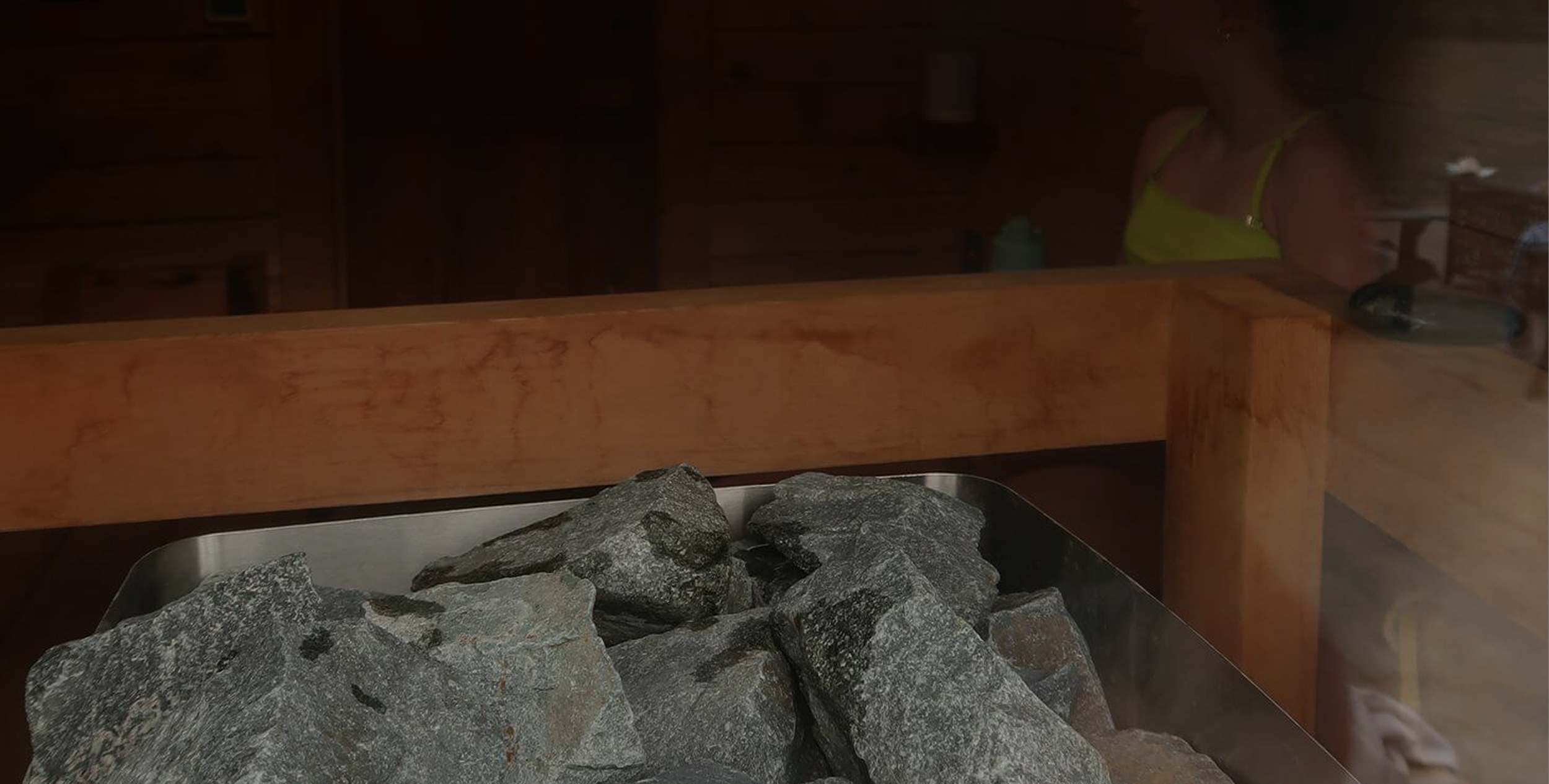 A sauna room with rocks inside a metal container, wooden benches along the walls, and a person in a bright yellow top visible through the glass window.
