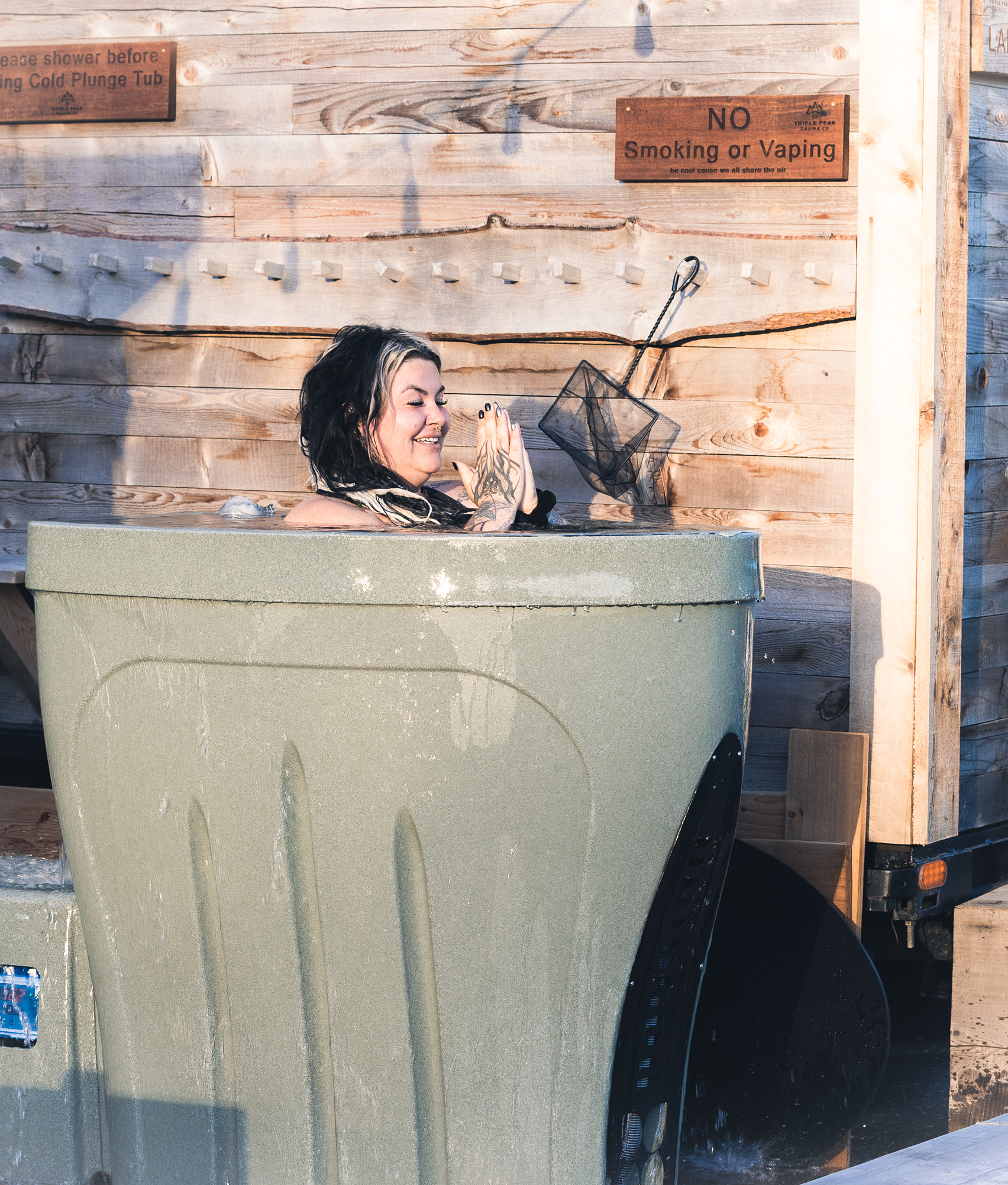 A woman with black and blonde hair enjoying a hot tub, with a wooden wall and signs indicating no smoking or vaping and shower rules in the background.