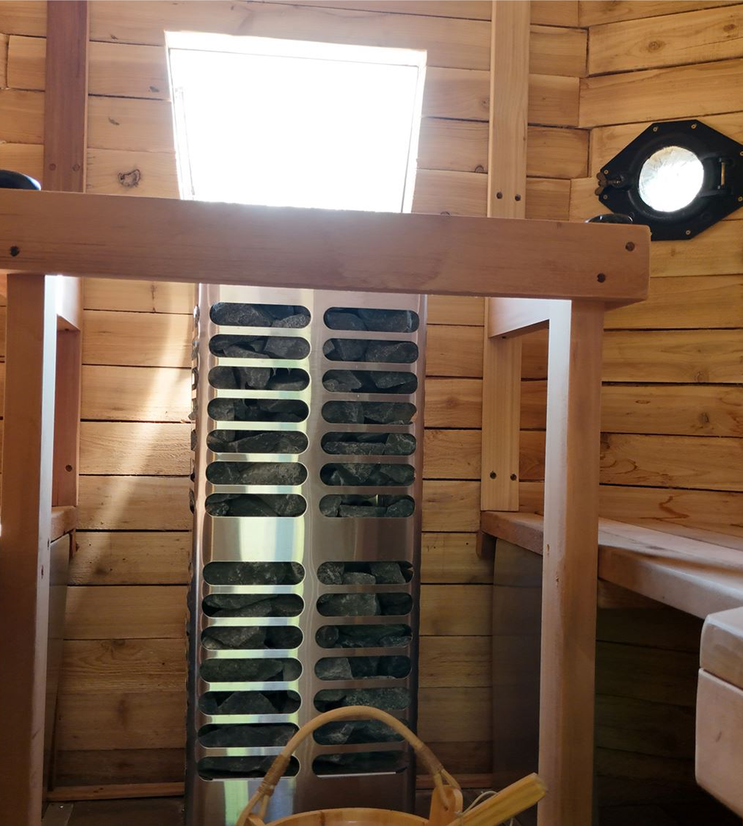 A wooden interior with a square window at the top, a ventilation vent on the right wall, a laundry basket filled with rocks, and a chair with a round back in the foreground.
