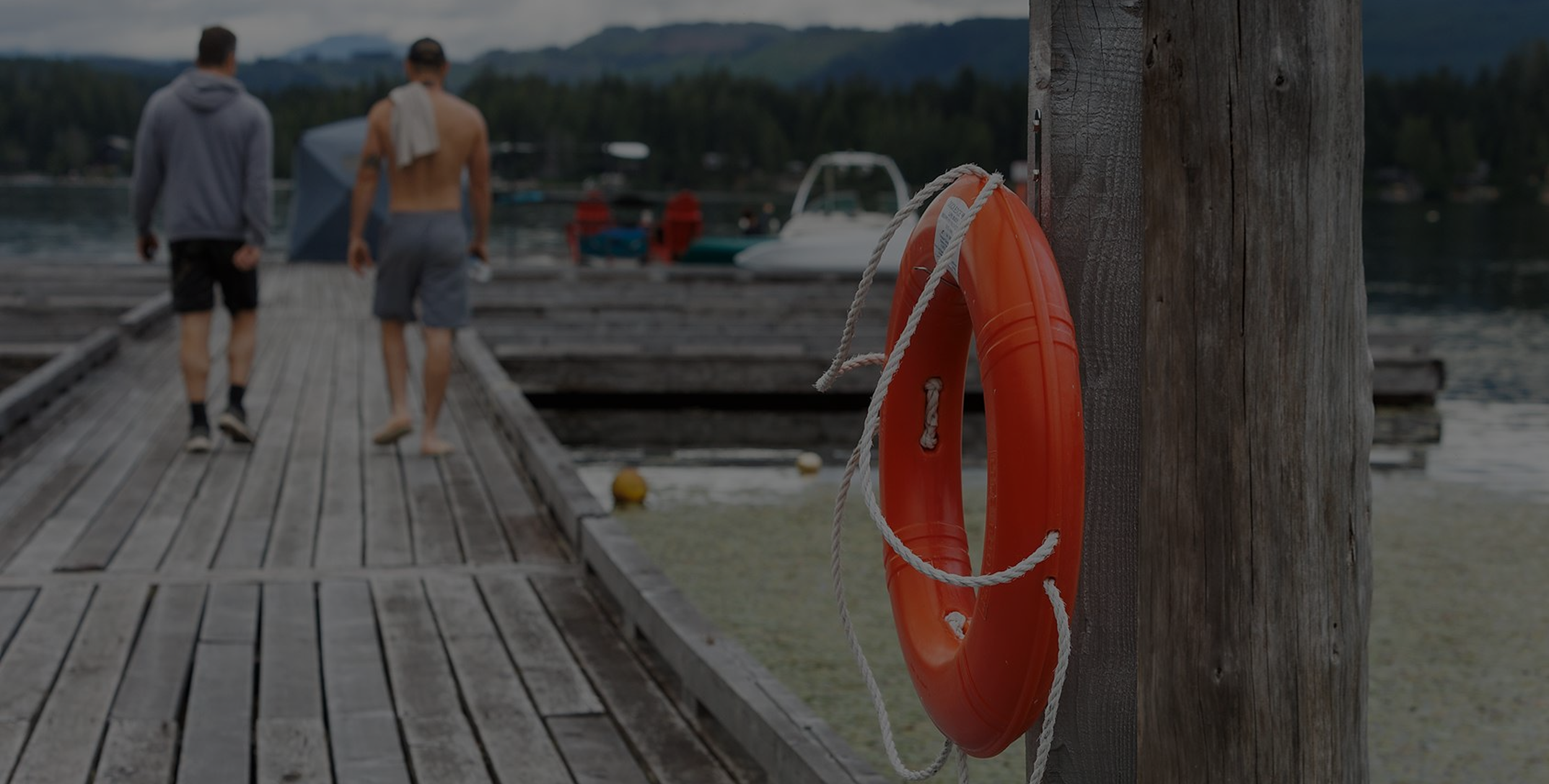 Two people walking on a wooden dock by a lake, with boats in the background and an orange life ring attached to a wooden post in the foreground.