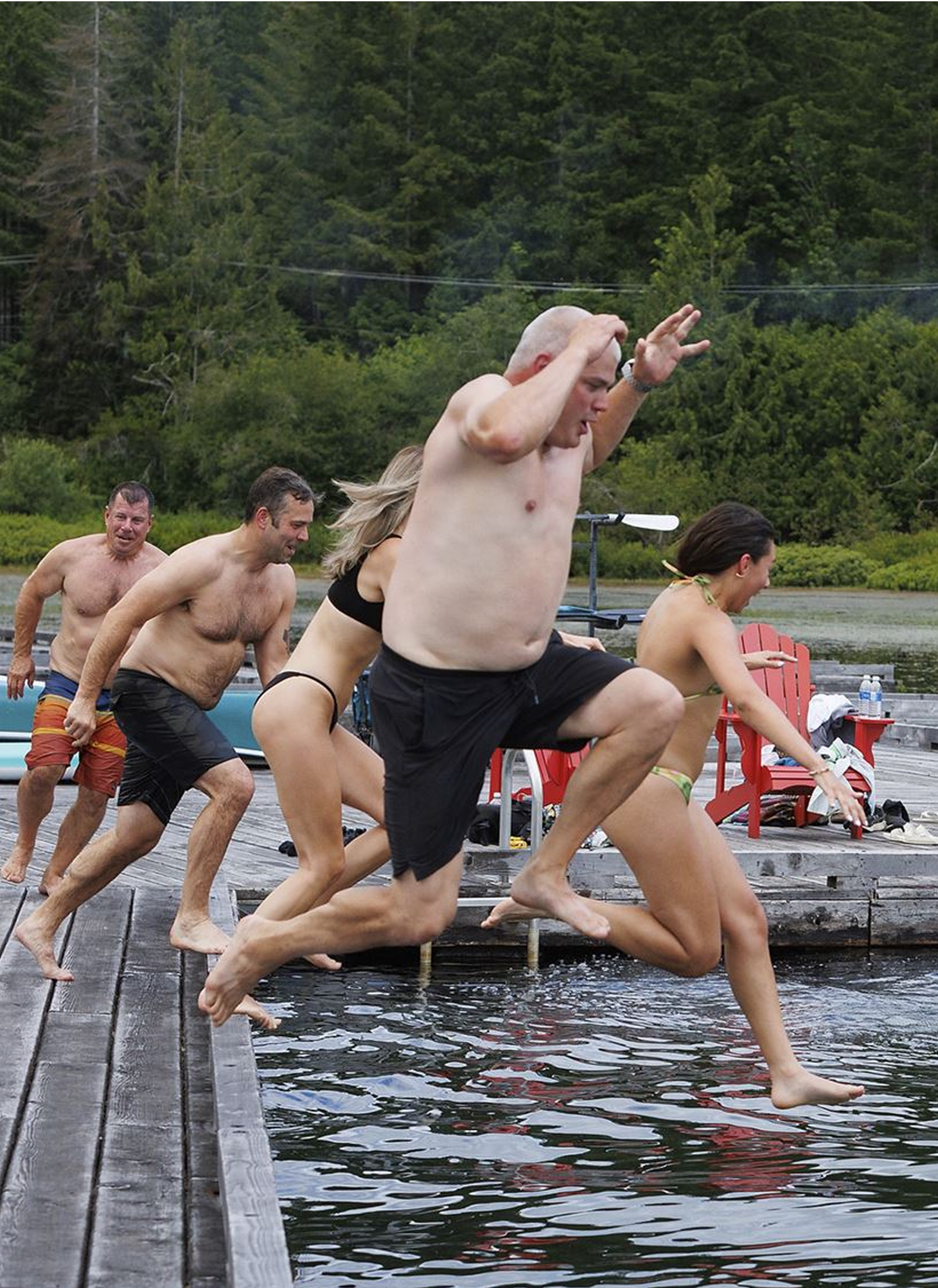 People jumping into a body of water from a dock, with green trees in the background.
