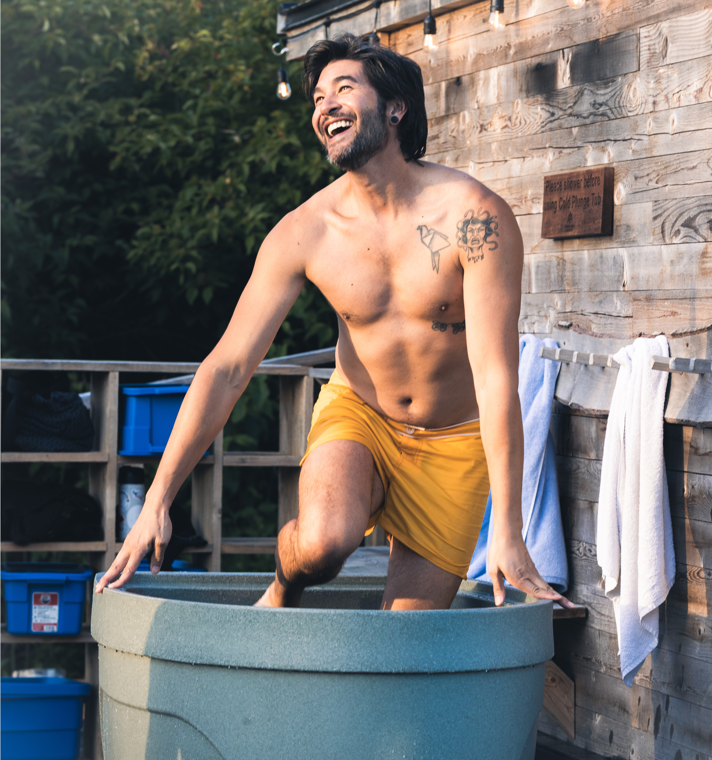 A shirtless man with a beard and tattoos, wearing yellow shorts, relaxing outdoors near a wooden wall with string lights, and a large blue plastic container in the foreground.