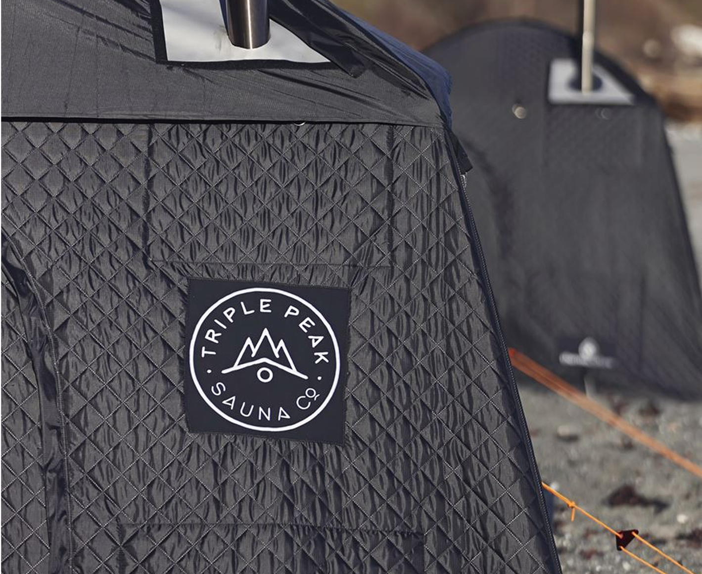 Close-up of a black tent with a Patch that reads 'TRIPLE PEAK SAUNA CO' and has a mountain logo, with additional tents and an orange guy line in the background.
