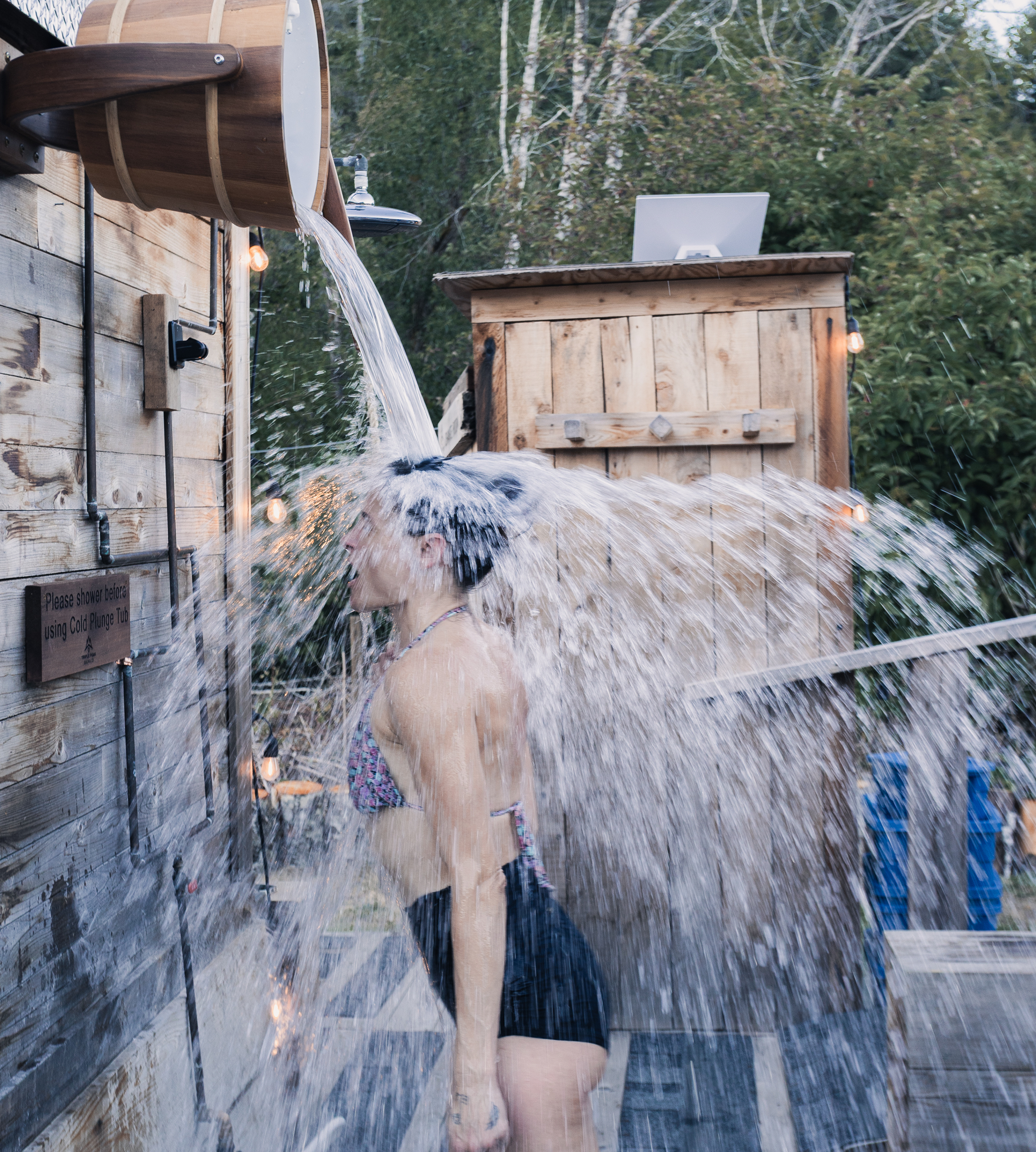 Person standing outside under a wooden shower with water pouring over their head and shoulders, surrounded by trees and wooden structures.
