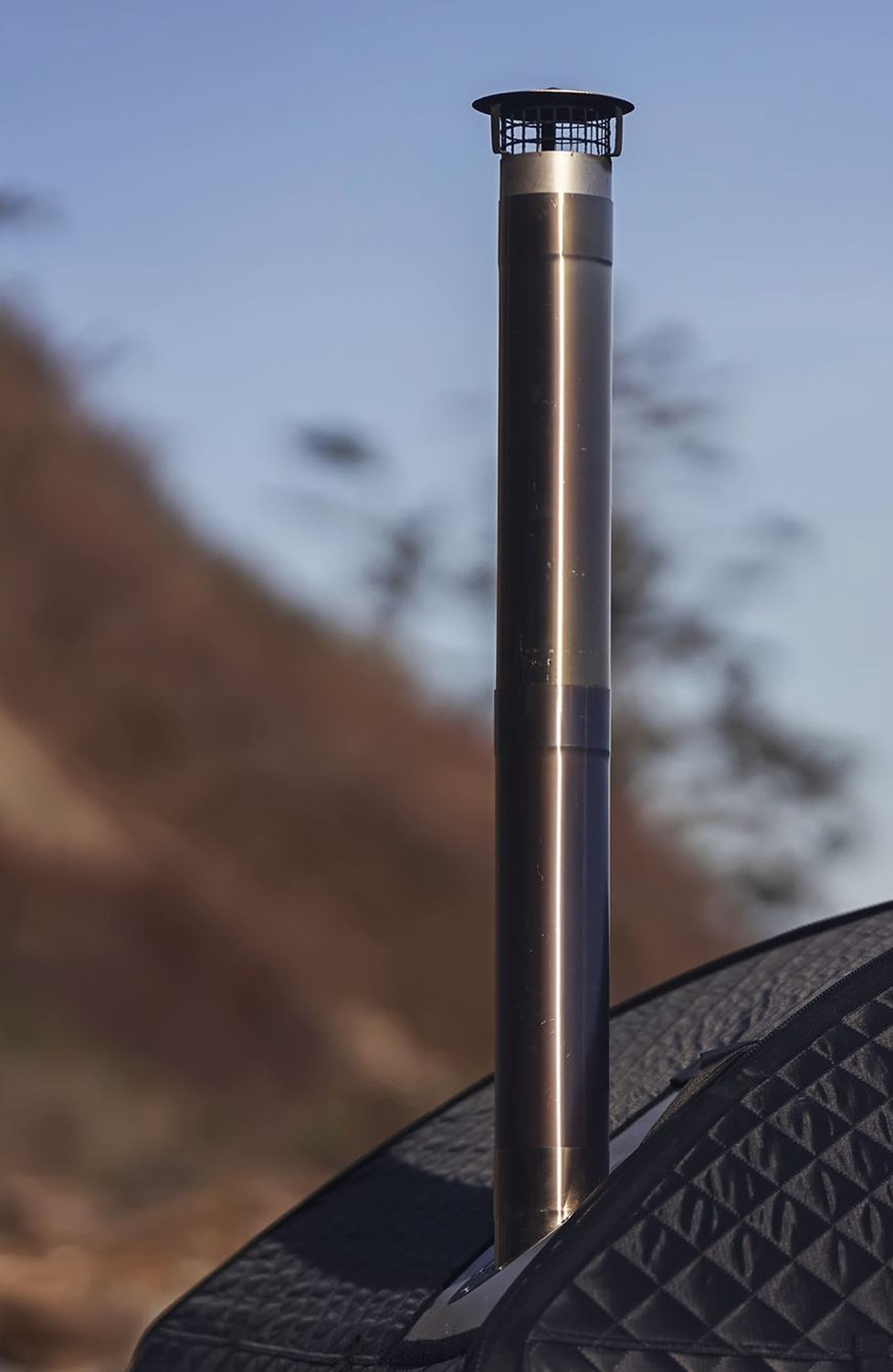 Close-up of a black golf bag with a metallic ventilation pipe rising from it, set outdoors with a blurred background of trees and sky.
