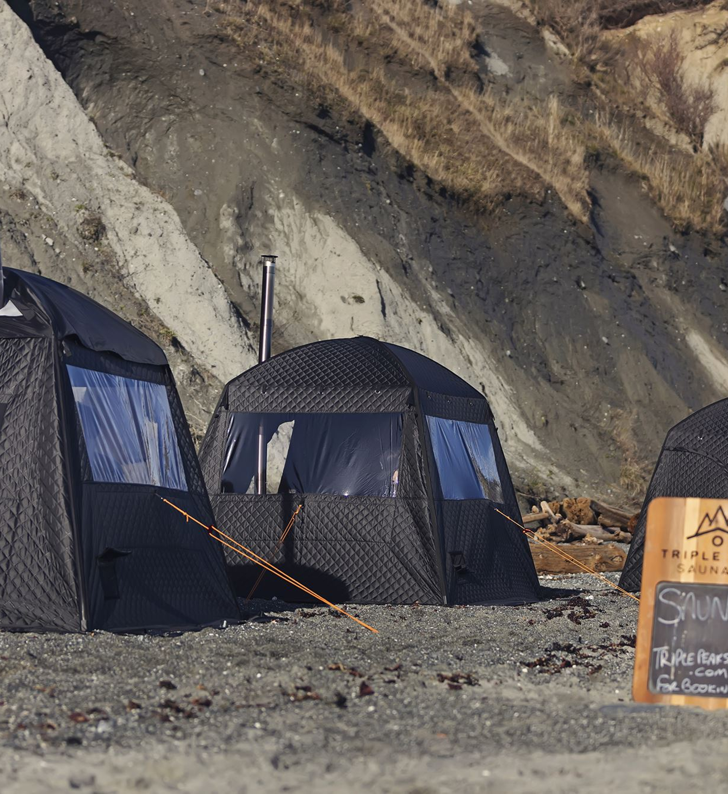 A camping site with black tents set up on dirt ground near rocky hillside, with a wooden sign on the right side of the image.