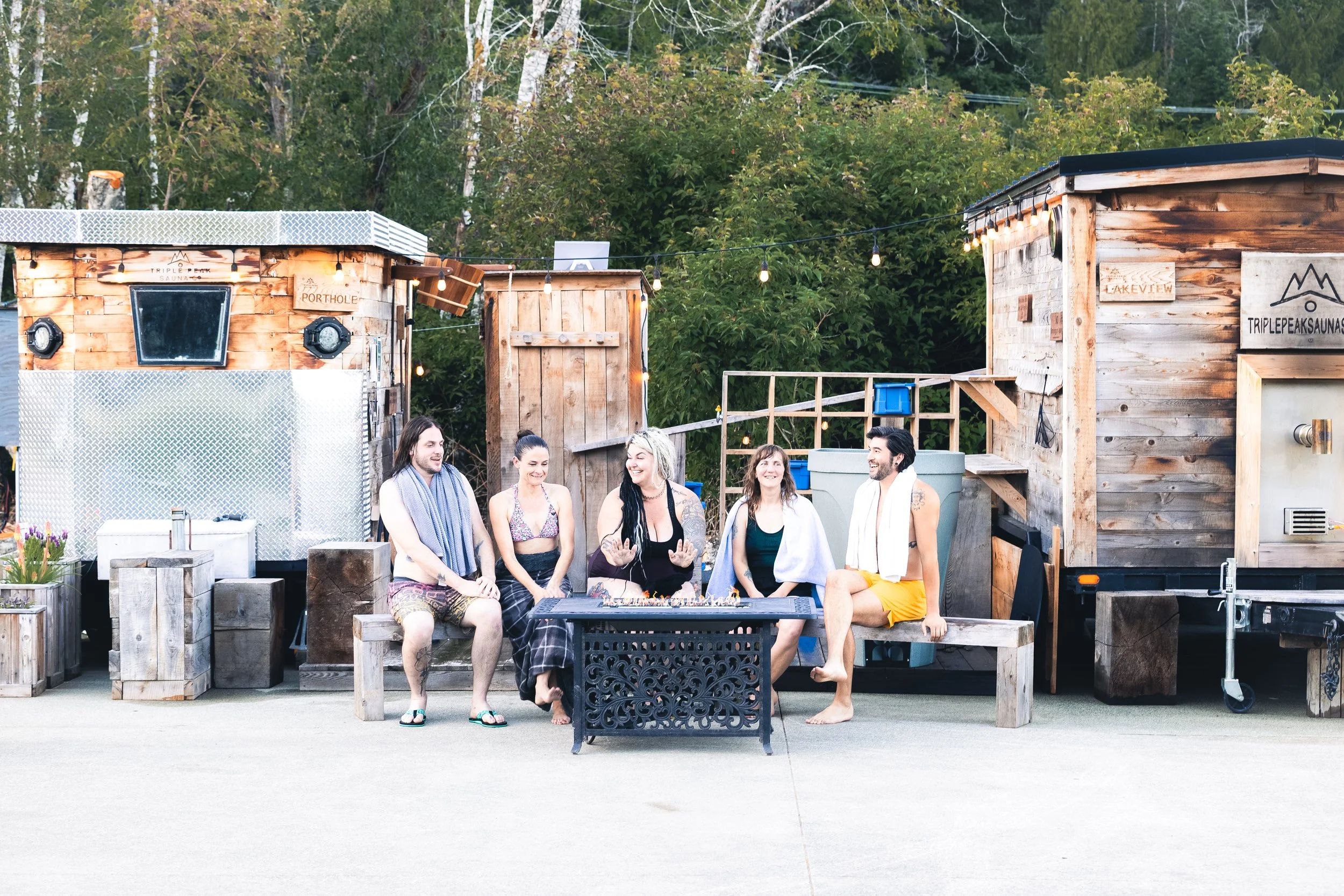 Group of five friends sitting around a fire pit in front of wooden structures with string lights, surrounded by trees, at an outdoor gathering.