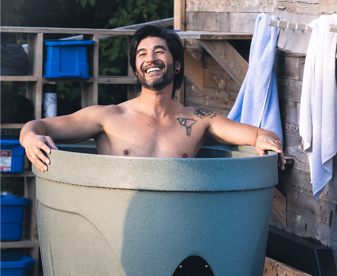 A smiling man with dark hair and tattoos relaxing in an outdoor hot tub with a wooden fence and shelves with blue storage bins and towels in the background.