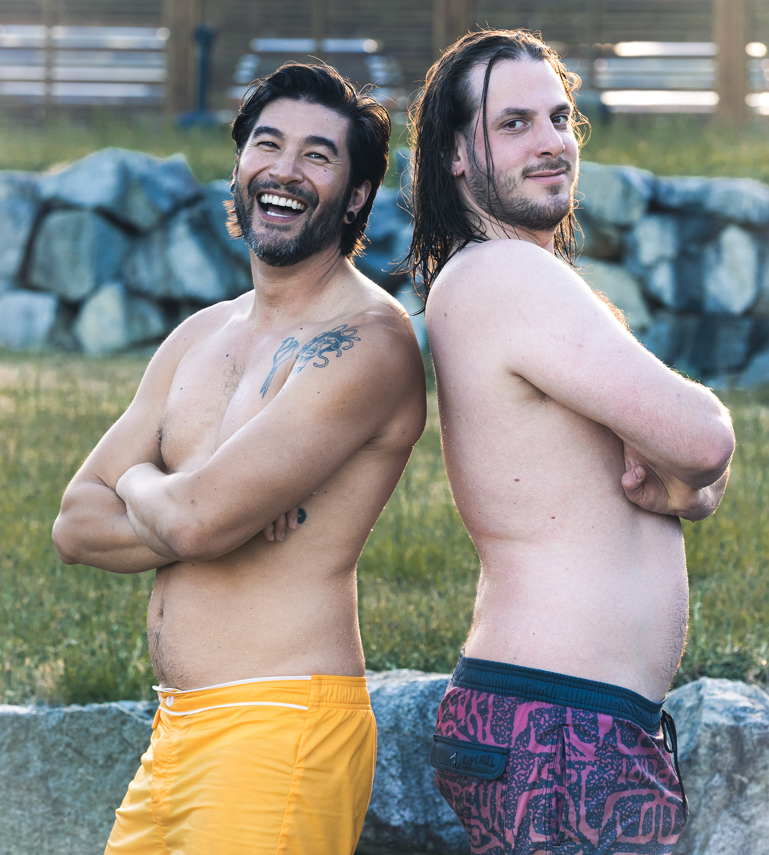 Two shirtless men standing back-to-back outdoors, smiling and looking at the camera, with a stone wall and a wooden fence in the background.