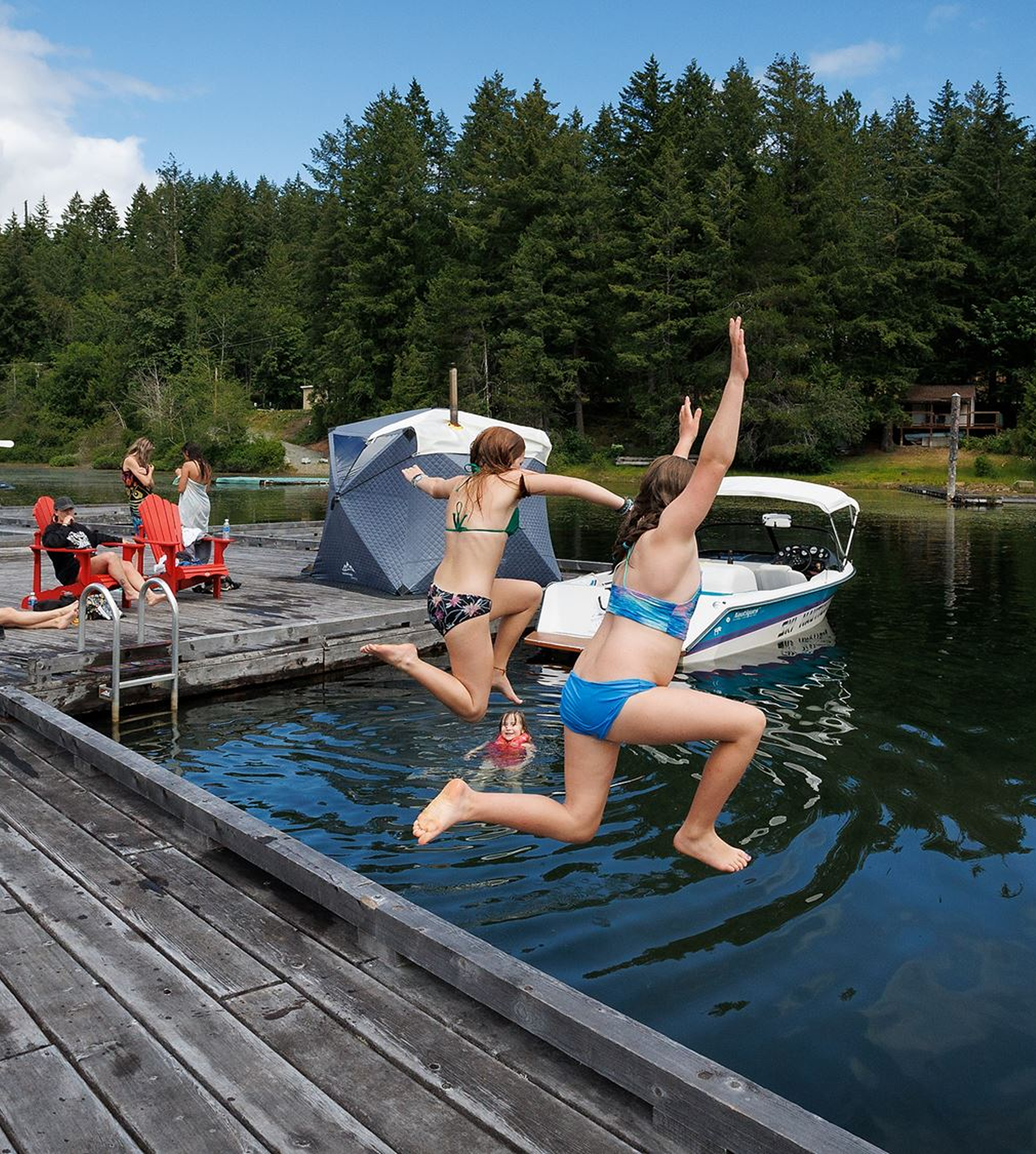 People enjoying a day by the lake with boats, a dock, and green trees in the background.