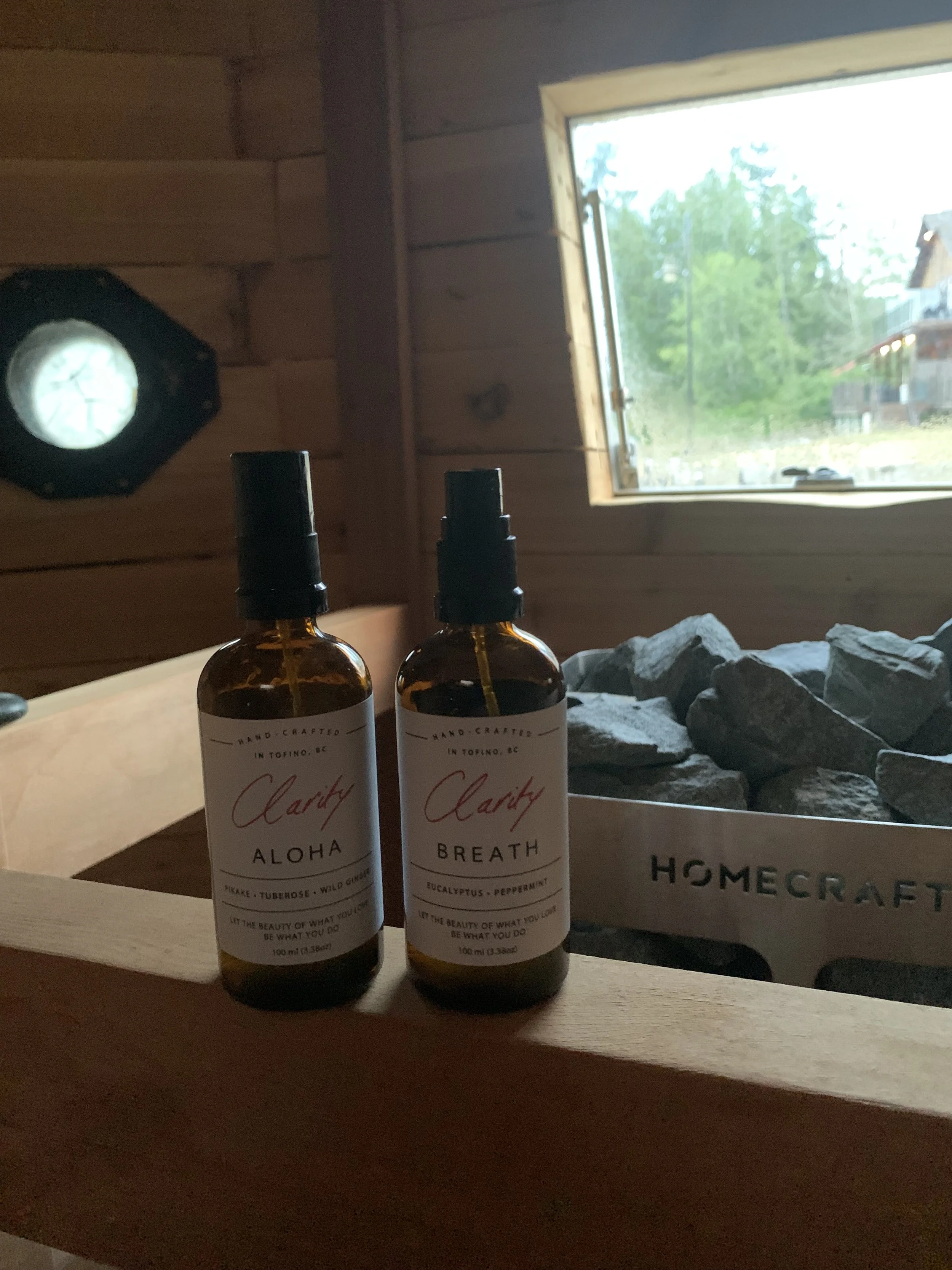 Two amber bottles with black spray caps placed on a wooden surface in front of a window, with a rock display behind them, and trees and houses visible outside the window.