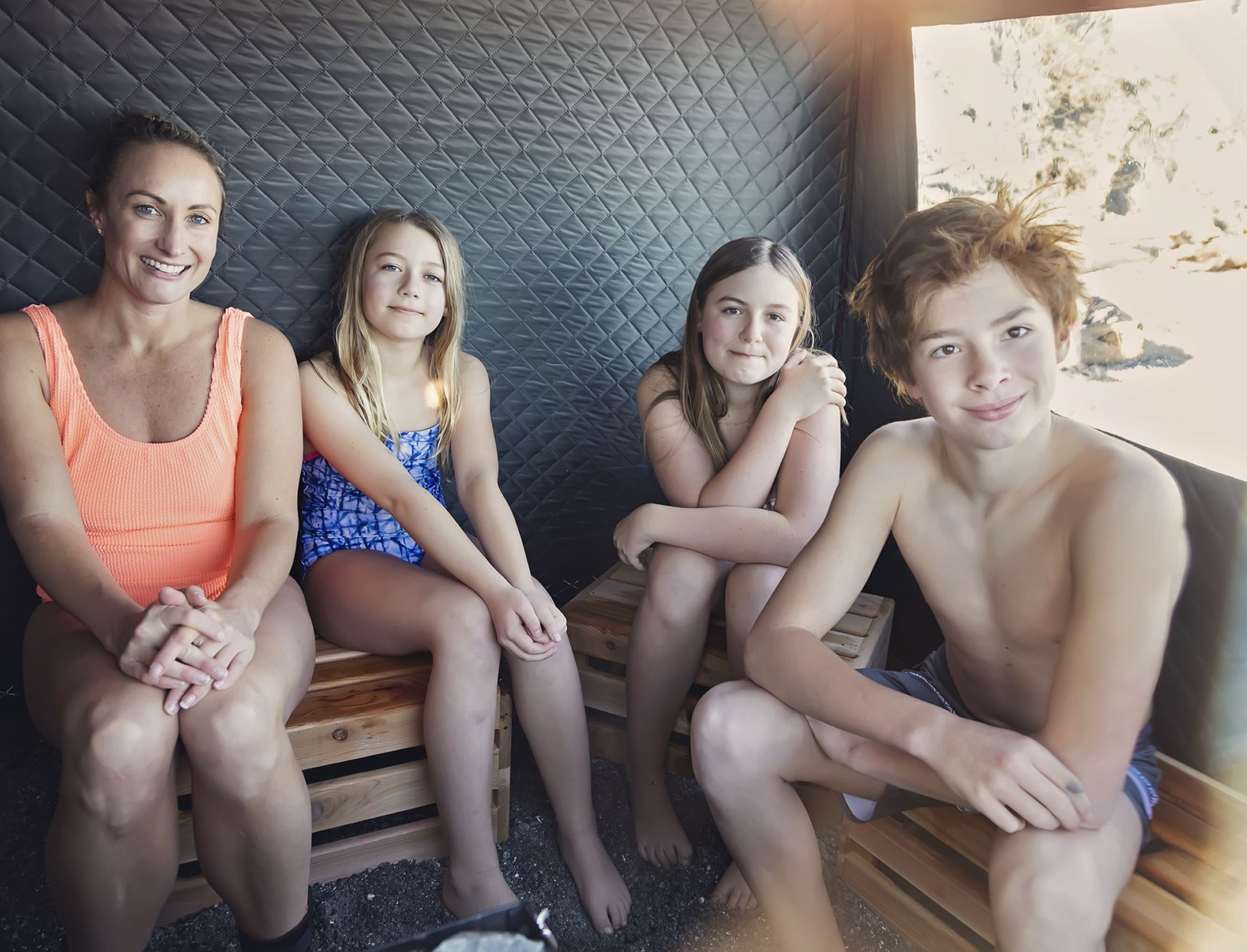 A group of five people, including a woman and four children, sitting together outside on a wooden bench, smiling and posing for the photo with a sunny sky and trees in the background.