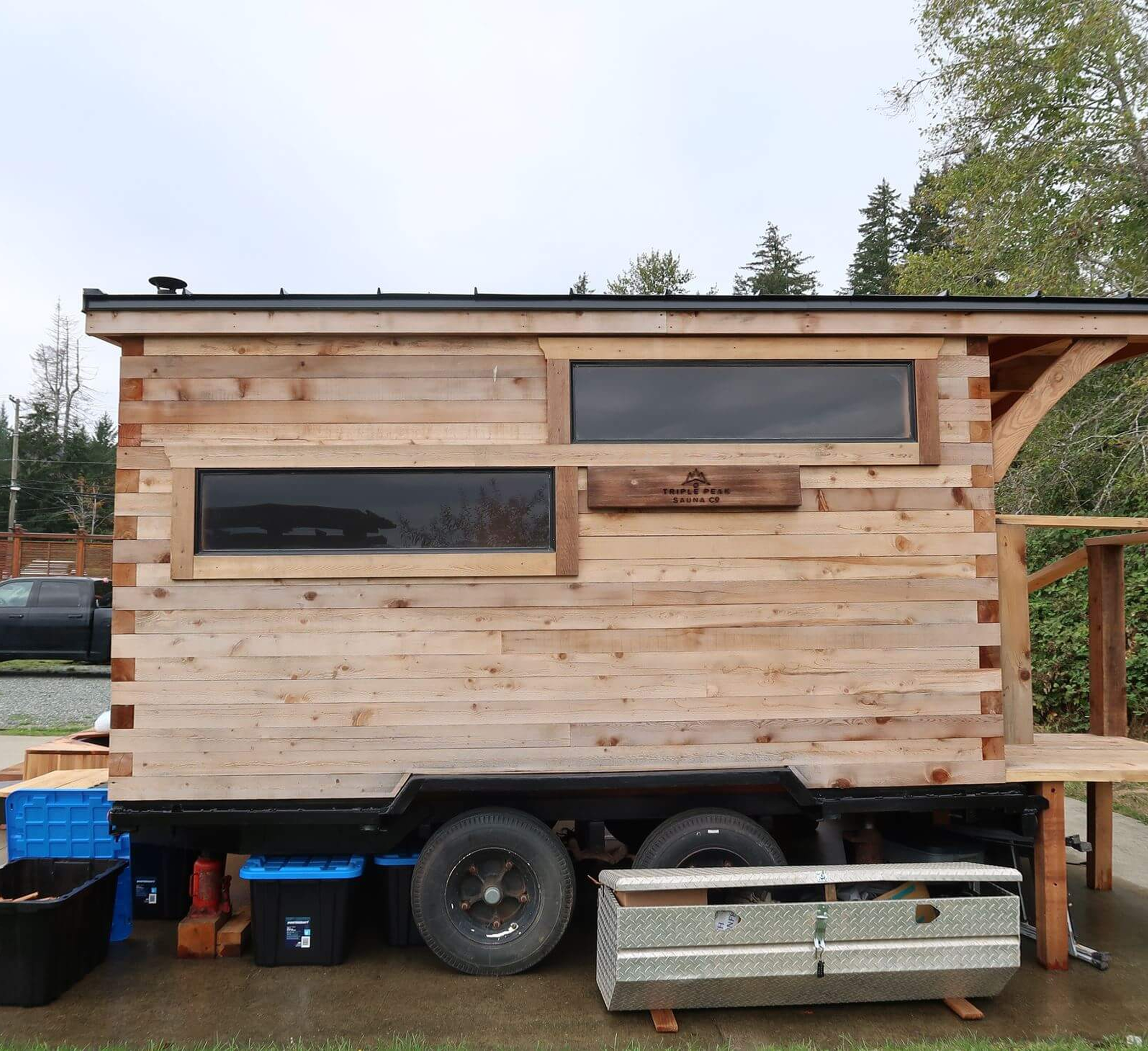 A tiny house under construction on a trailer with exposed wooden siding, dual large windows, and a sign that reads 'Triple Peak Sauna Co.' in a wooded outdoor area.
