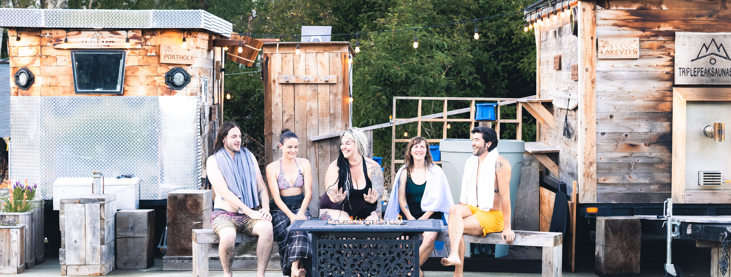 A group of six friends sitting around a fire pit, enjoying each other's company outside in a backyard with wooden structures and string lights.