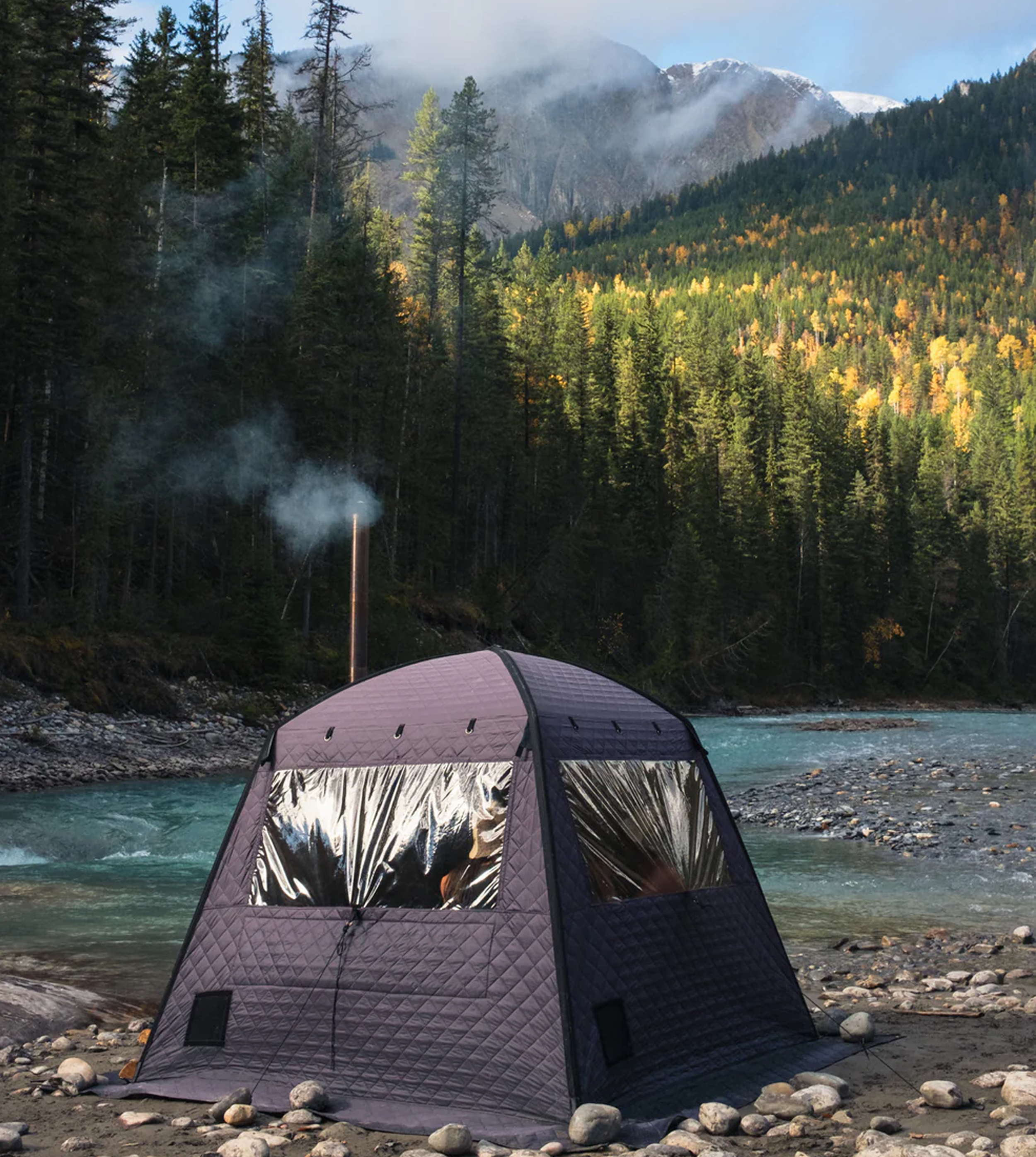 A camping tent set up on a rocky riverbank with a forested mountainside in the background, snow-capped peaks, and a partly cloudy sky.