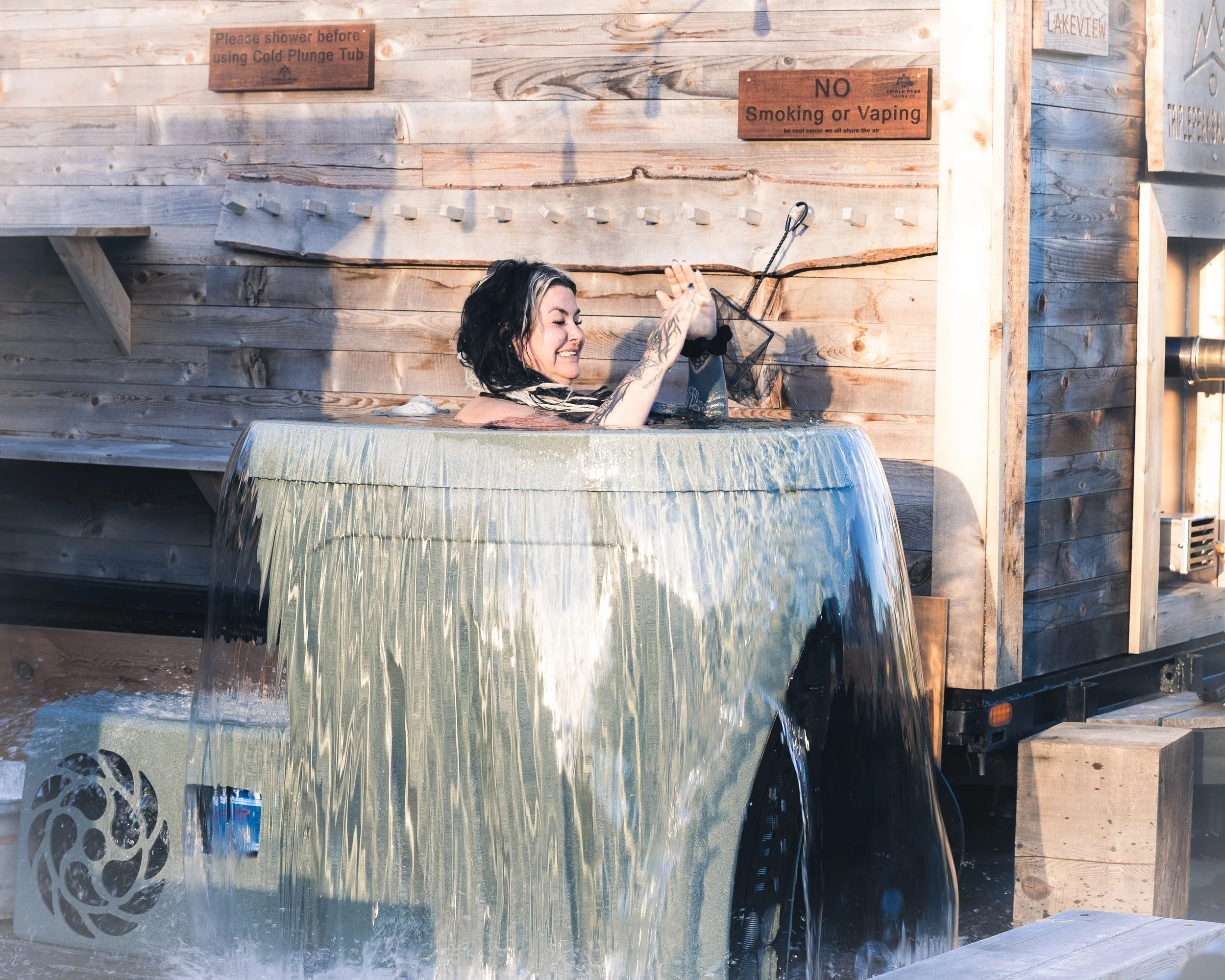 A woman with tattoos and black and blond hair enjoying a soak in an outdoor spillway hot tub with water cascading down a metal wall, wooden wall with signs, and textured ground, located at a rustic outdoor setting.