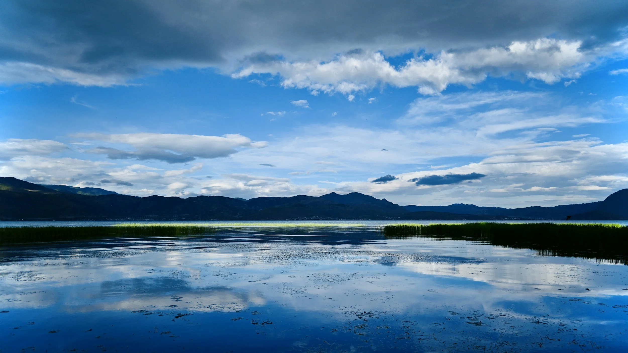 Scenic landscape with a large lake reflecting the cloudy sky and mountains in the background.