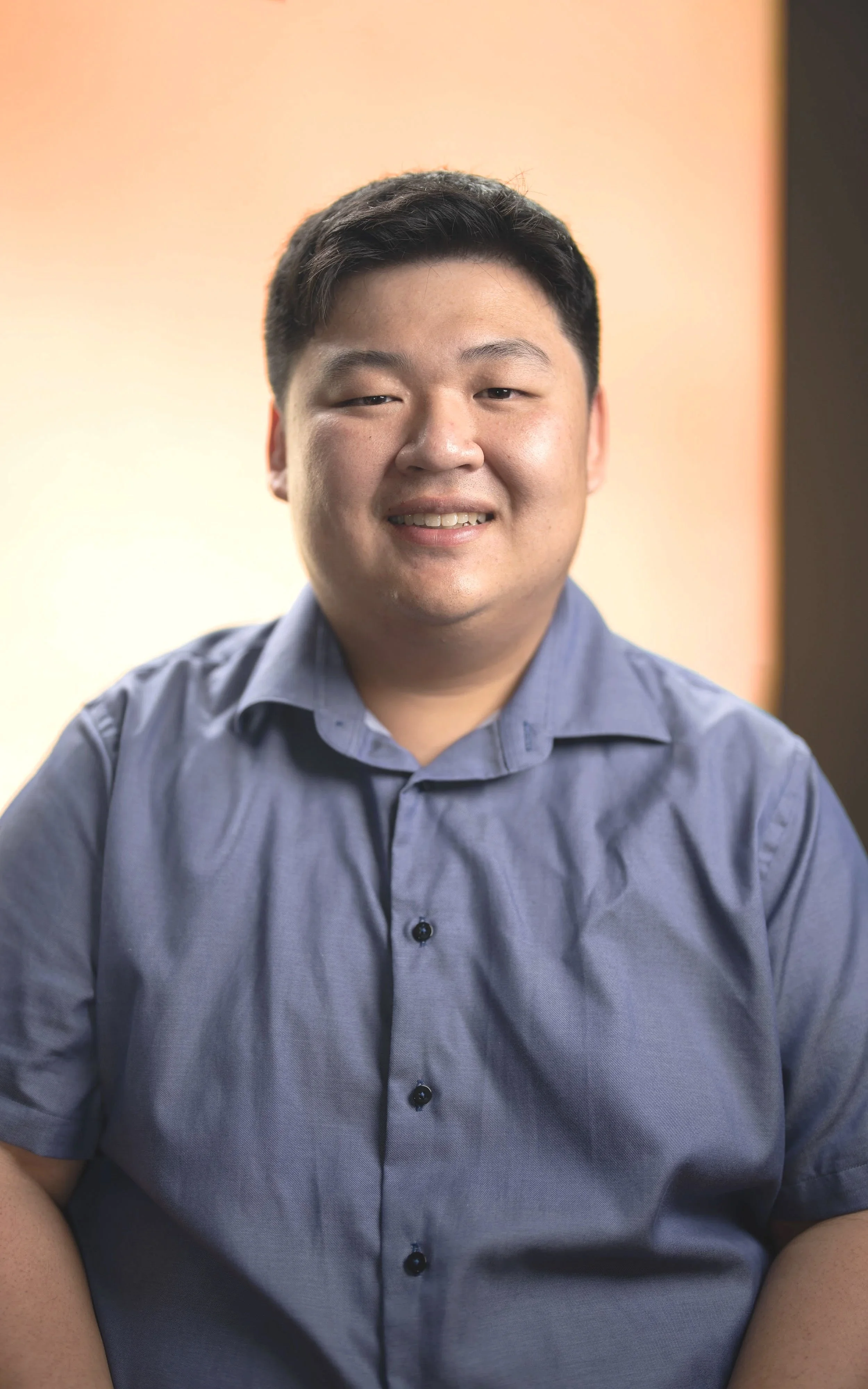 A young man with short black hair wearing a light blue button-up shirt smiling at the camera. The background features a gradient of warm colors from light pink to beige.