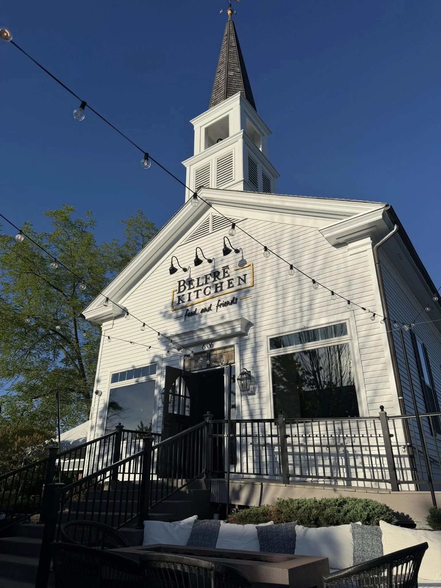 Exterior of a white church with a tall steeple, string lights hanging across the courtyard, steps leading up to the entrance, and outdoor seating with cushions in the foreground. Clear blue sky with some trees.