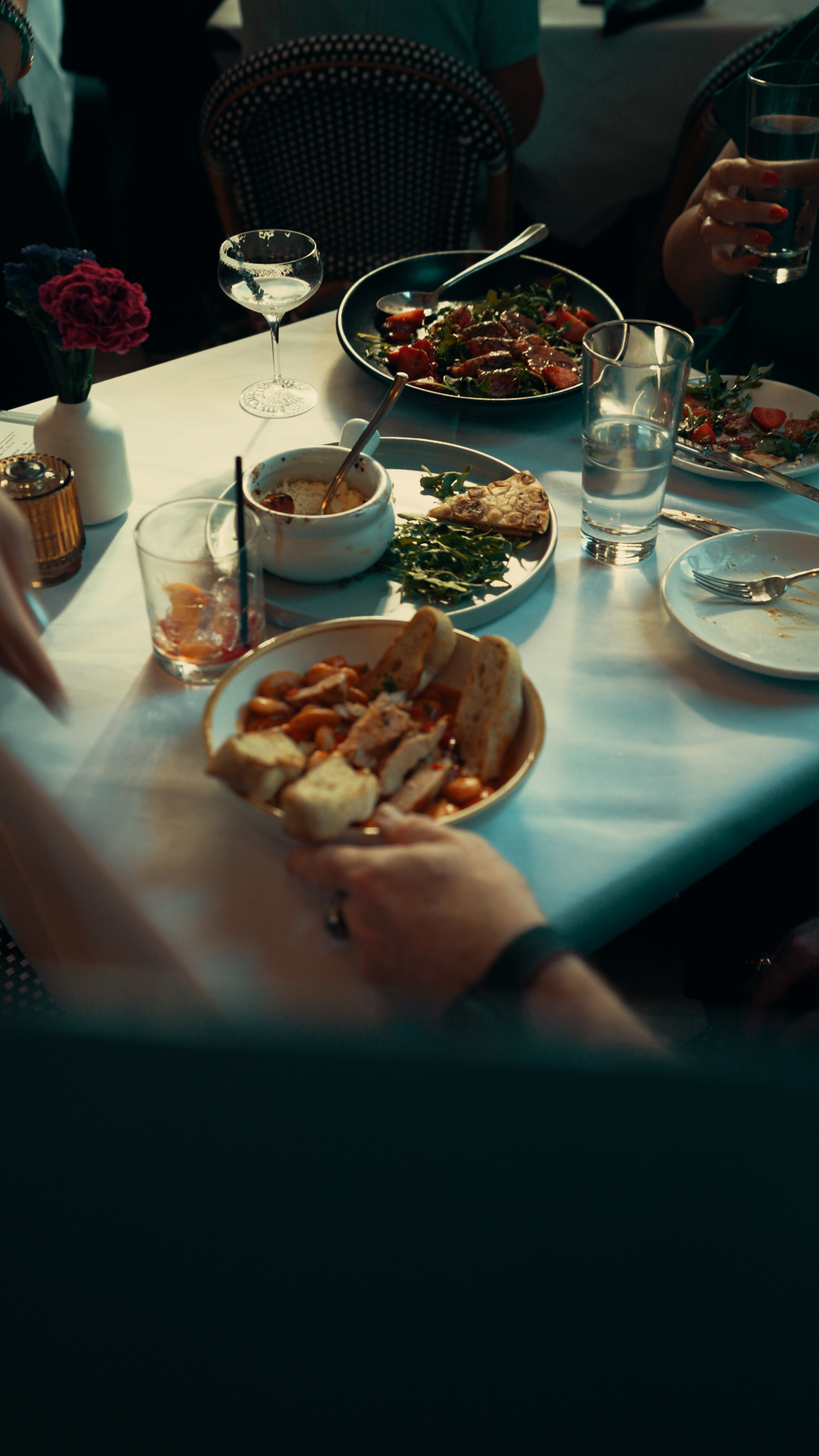 A dinner table with plates of food, including bread, salad, and dessert, along with glasses of water and wine, and a small vase with pink and purple flowers.