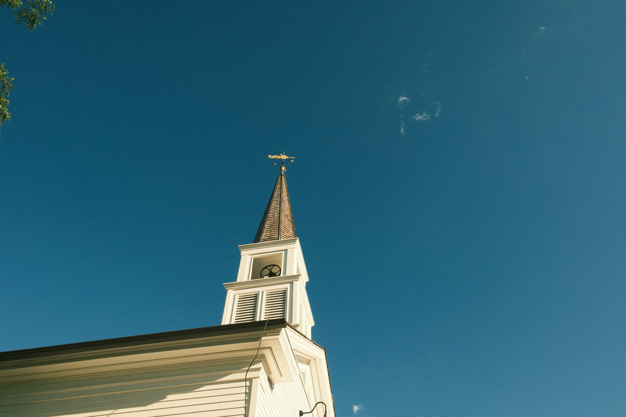 The steeple of a white church with a weather vane on top, set against a clear blue sky with a few small clouds.