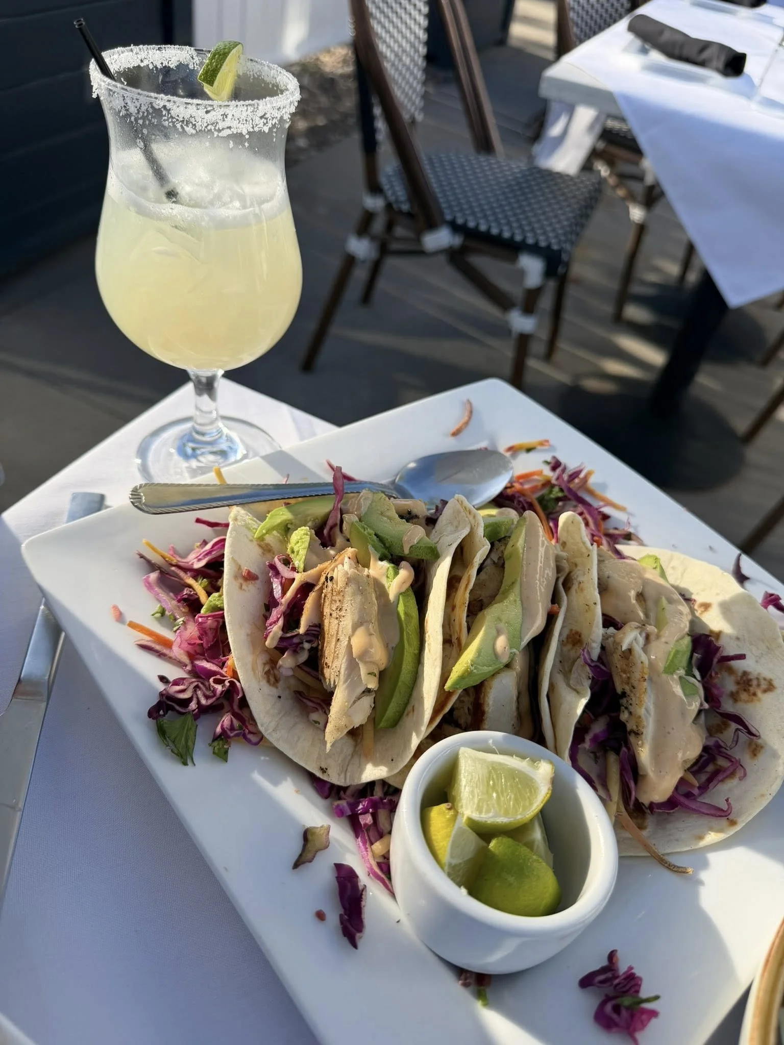 Plate of tacos filled with meat, avocado, and vegetables, topped with shredded cabbage and served with lime wedges and a small bowl of lime slices, on a white table outdoor setting.