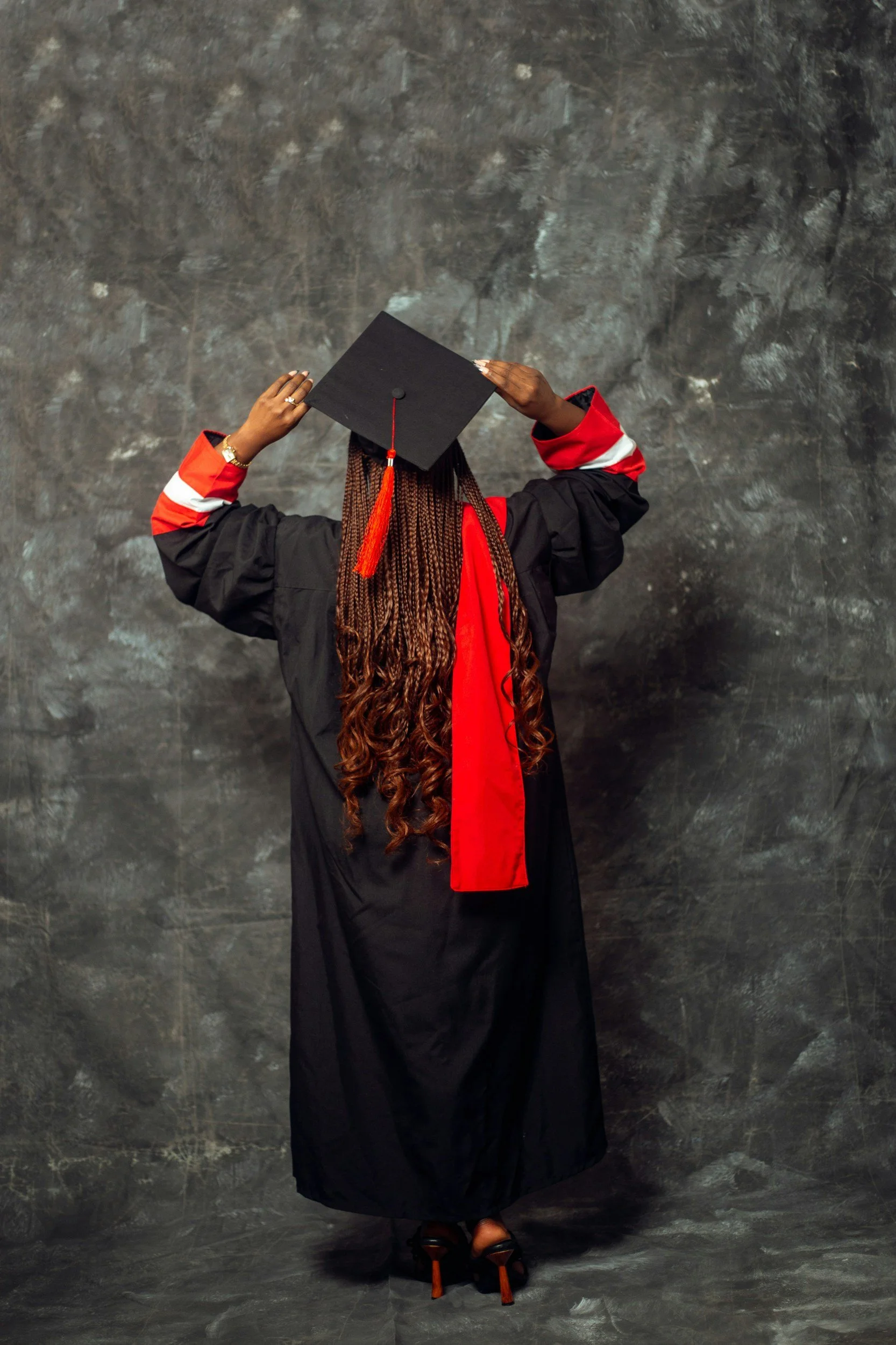 A woman in a graduation cap and gown standing against a gray textured background, holding her cap with both hands.