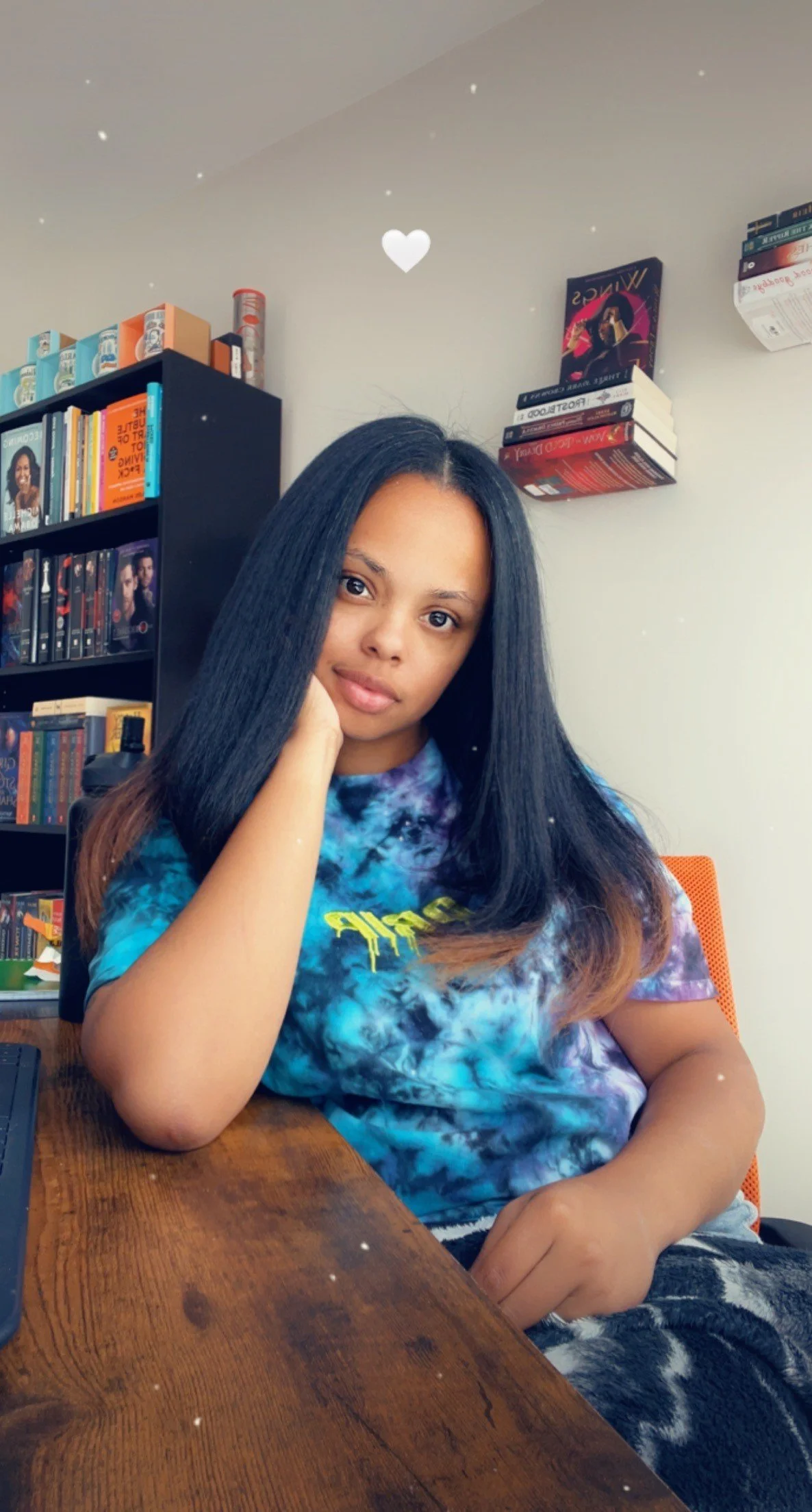 A young woman with long black hair sitting at a wooden table, resting her head on her hand, in a room with shelves of books and DVDs in the background.