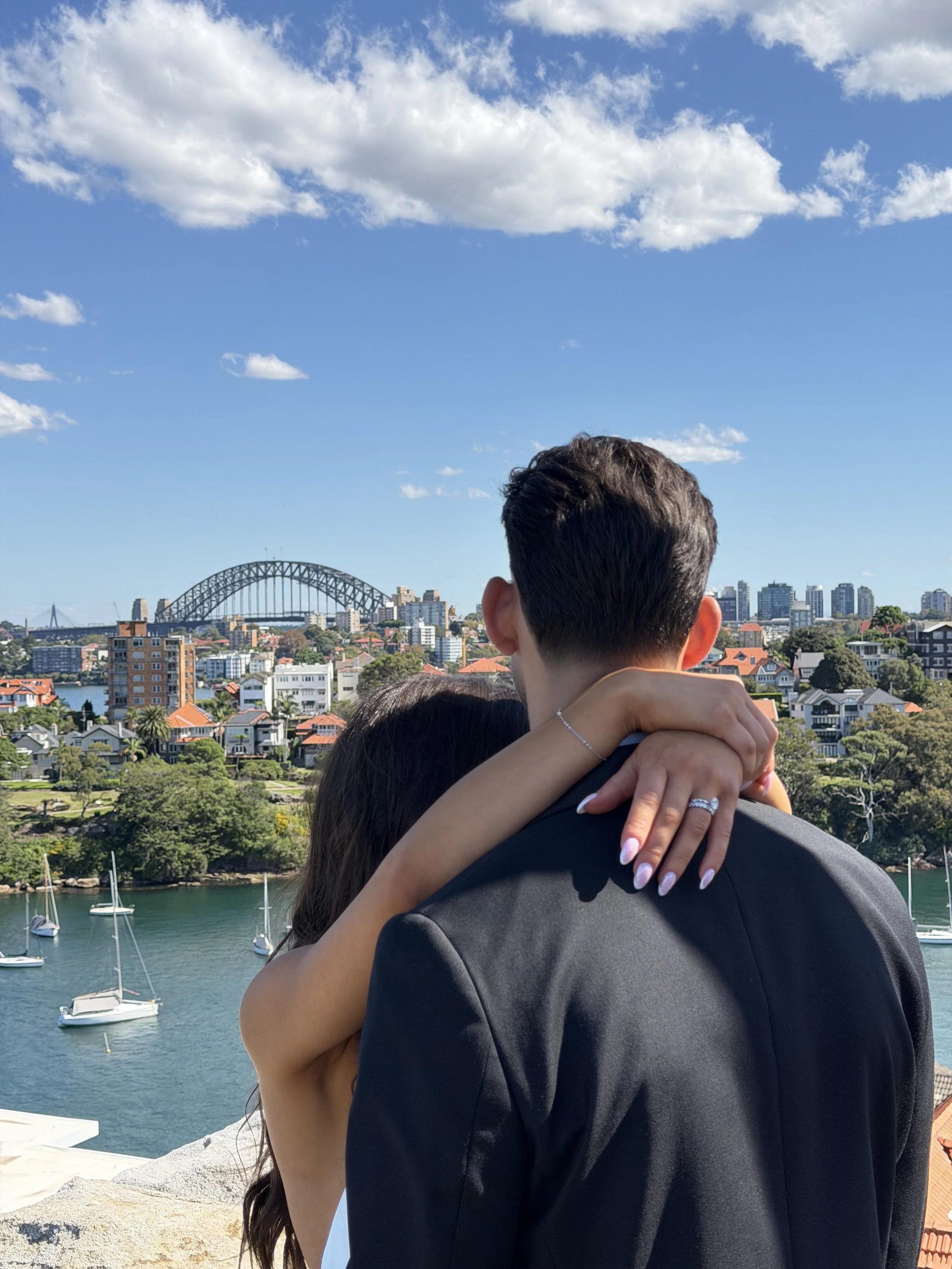 A couple hugging outdoors near a waterfront with boats docked in the water. The man is wearing a gray suit, and the woman has long dark hair and is wearing a silver bracelet and ring. In the background, there are city buildings, a bridge, and a blue sky with scattered clouds.