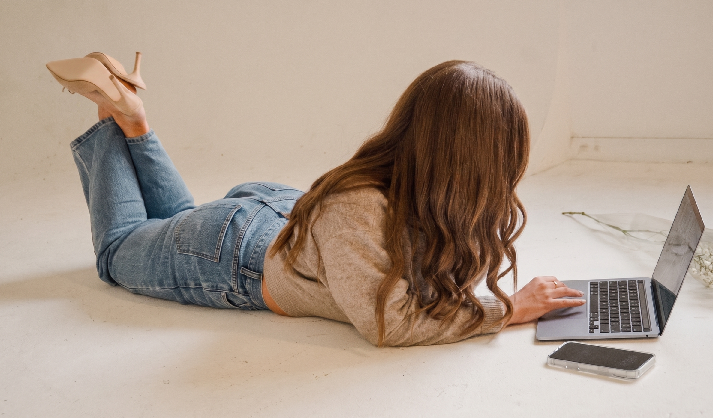 A woman with long wavy hair lying on her stomach on a light-colored floor, working on a laptop. She is wearing blue jeans and a beige sweater. There is a phone and a glass container with white flowers nearby. The background is plain and light-colored.