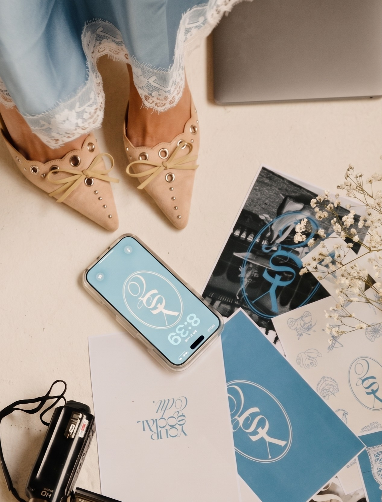 Pointed beige shoes with decorative eyelets, paired with lace-trimmed light blue jeans, placed on a white surface alongside printed materials, a smartphone with a logo, and a vintage camera.