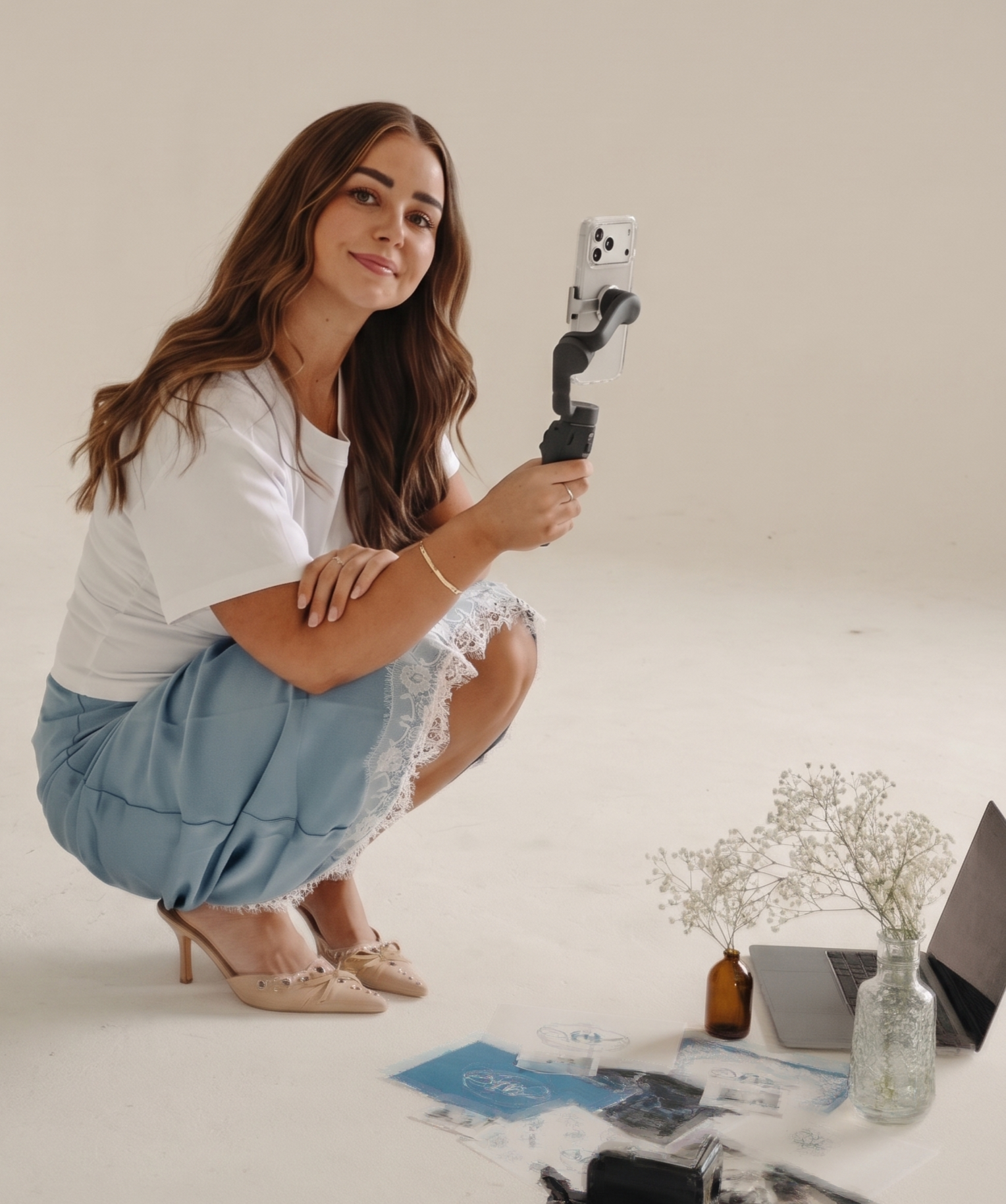 A woman with long wavy brown hair taking a selfie with a smartphone on a gimbal. She is wearing a white top, a blue skirt, and beige high heels. She is crouching on a white floor next to a small table with a laptop, a brown vase with flowers, a glass jar, and some papers.