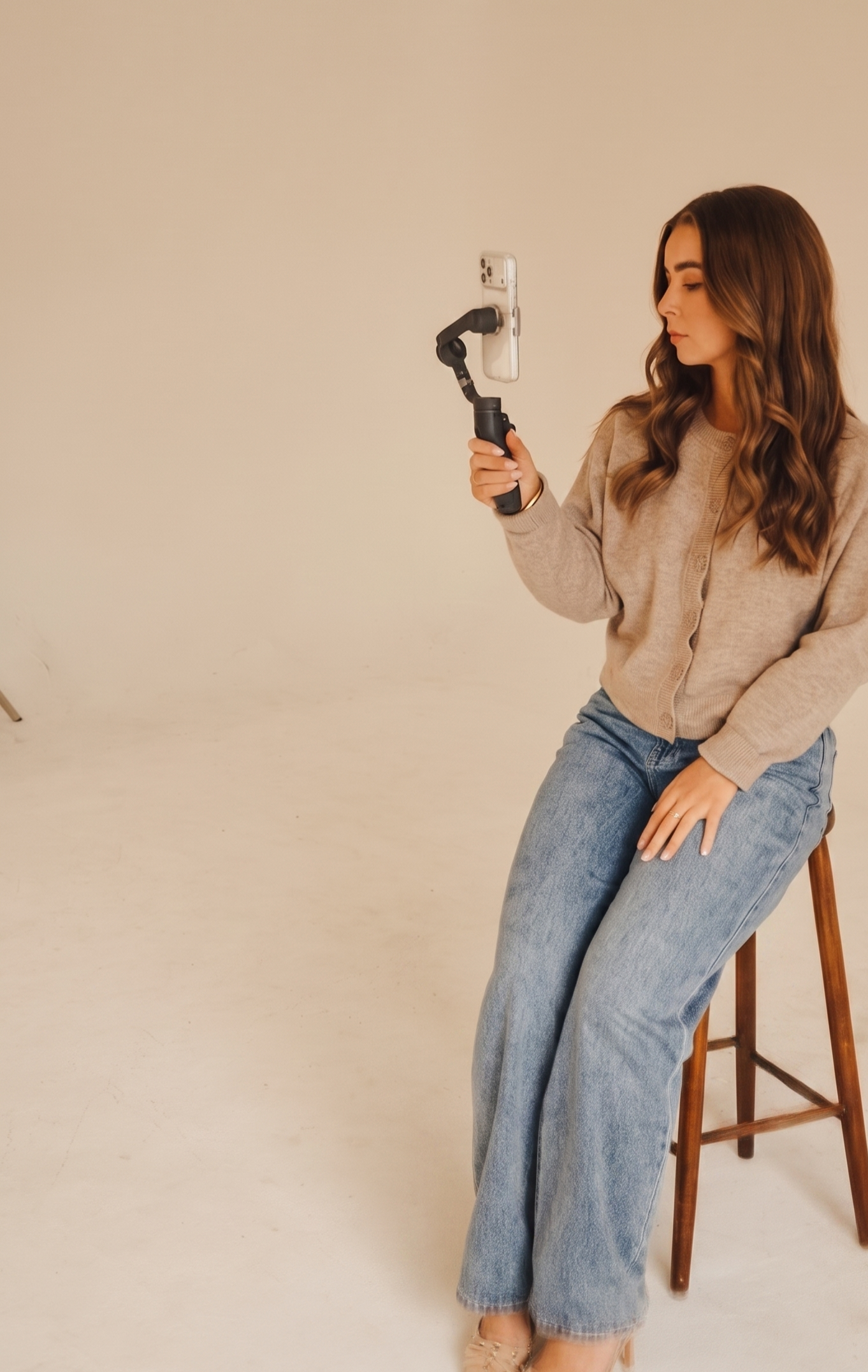 A woman with long wavy brown hair, wearing a beige sweater and light-washed jeans, is sitting on a stool and taking a selfie with a smartphone mounted on a handheld device in a plain white room.