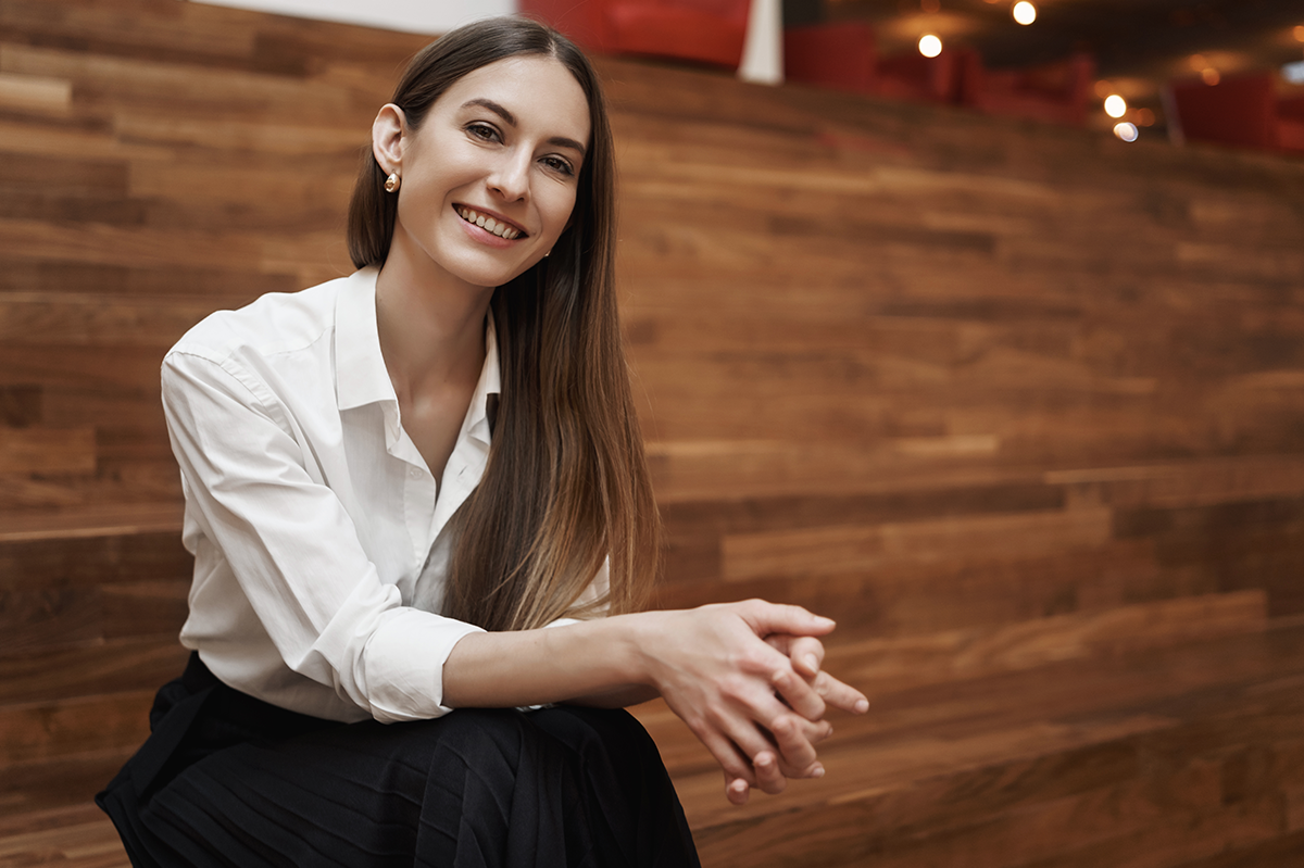 Mujer joven sonriendo, vestida con blusa blanca y falda negra, sentada en un entorno de madera con luces cálidas en el fondo.