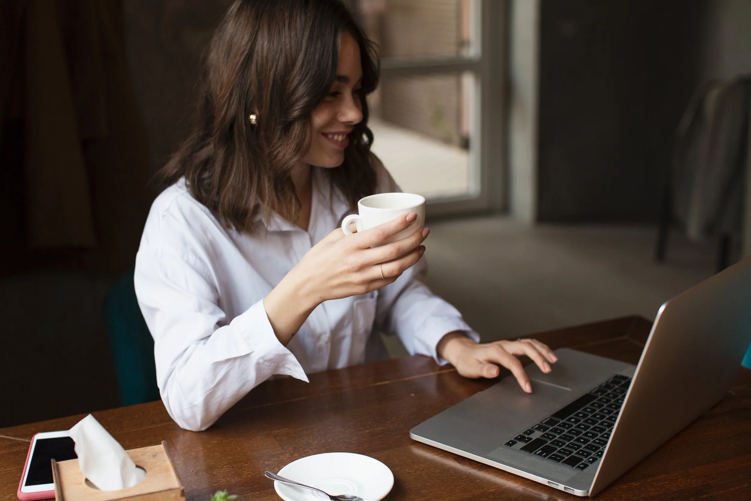 Una mujer sonriendo mientras toma una taza de café y trabaja en una laptop en una mesa de madera en un ambiente interior.