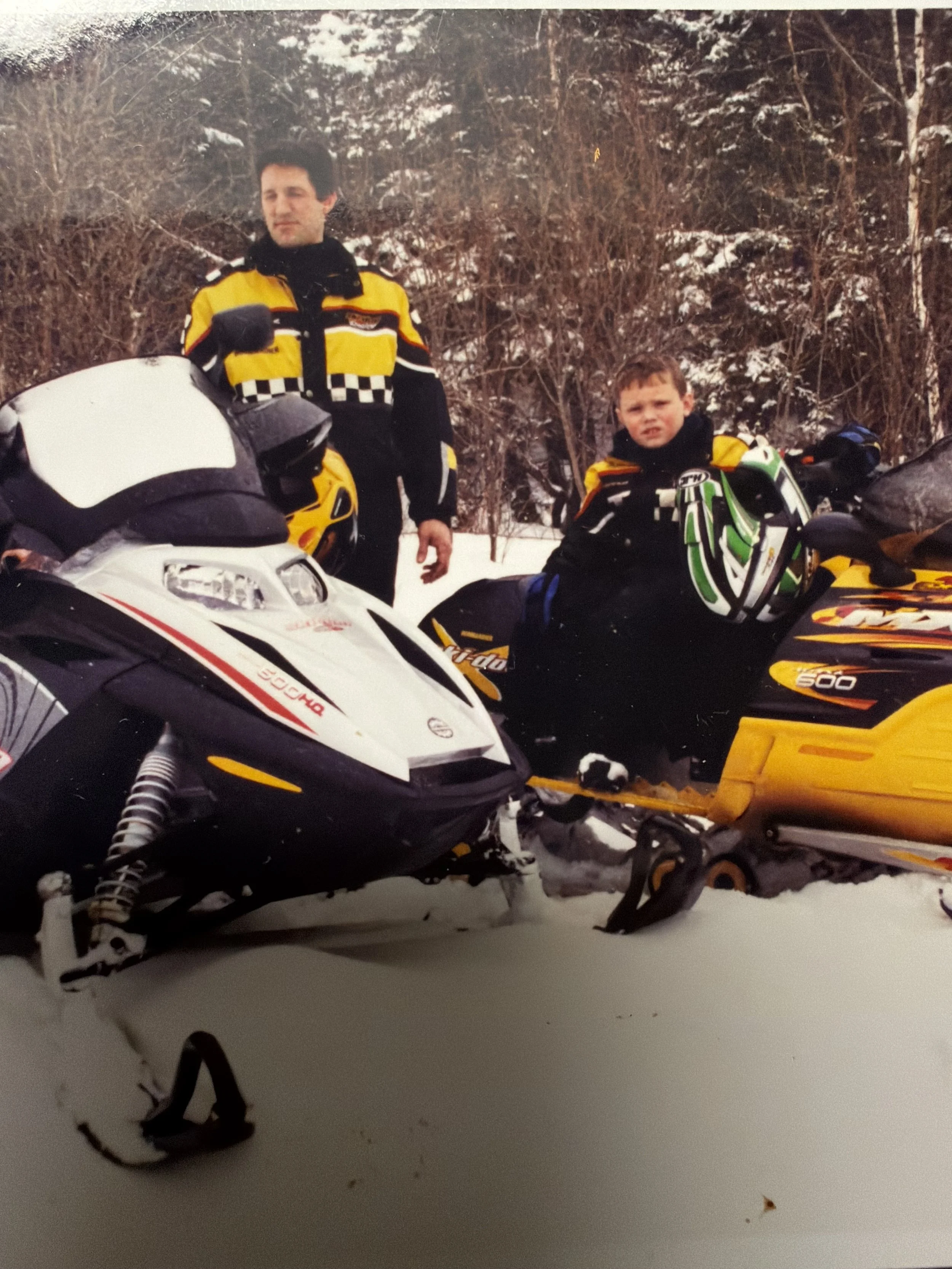 Two men in motorcycle gear standing outdoors in a snowy environment with trees and snow-covered ground, with snowmobiles nearby.
