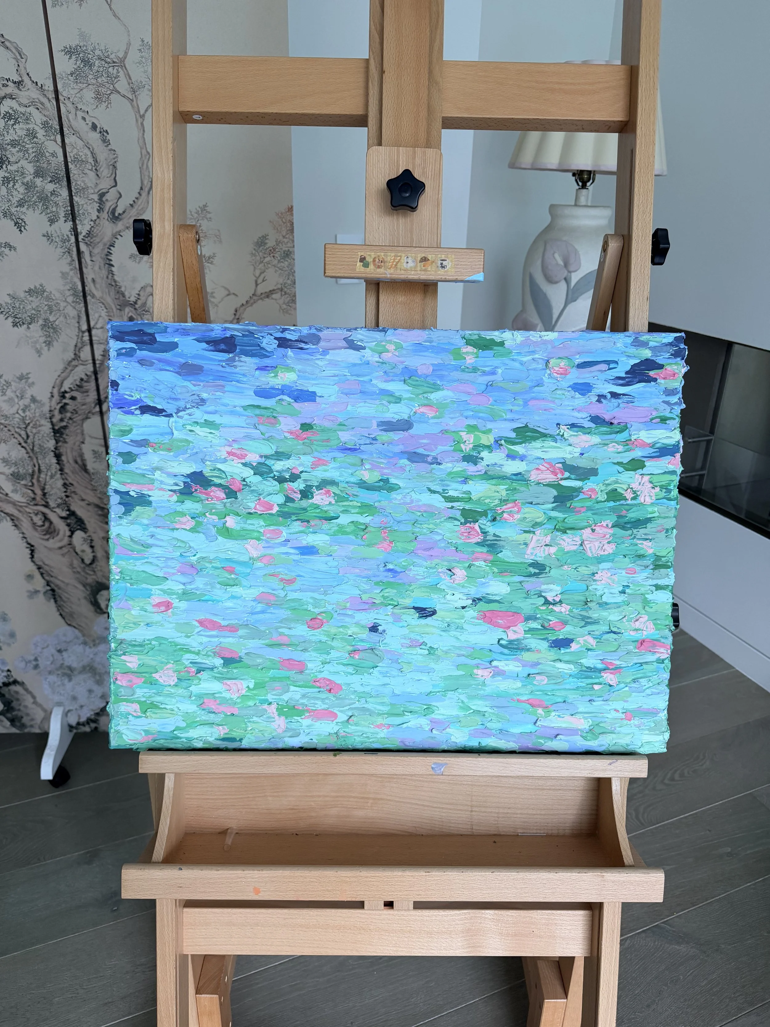 A painting on a canvas with textured brushstrokes in shades of blue, green, pink, and lavender, sitting on a wooden easel in a room with decorative wallpaper and a white ceramic lamp.