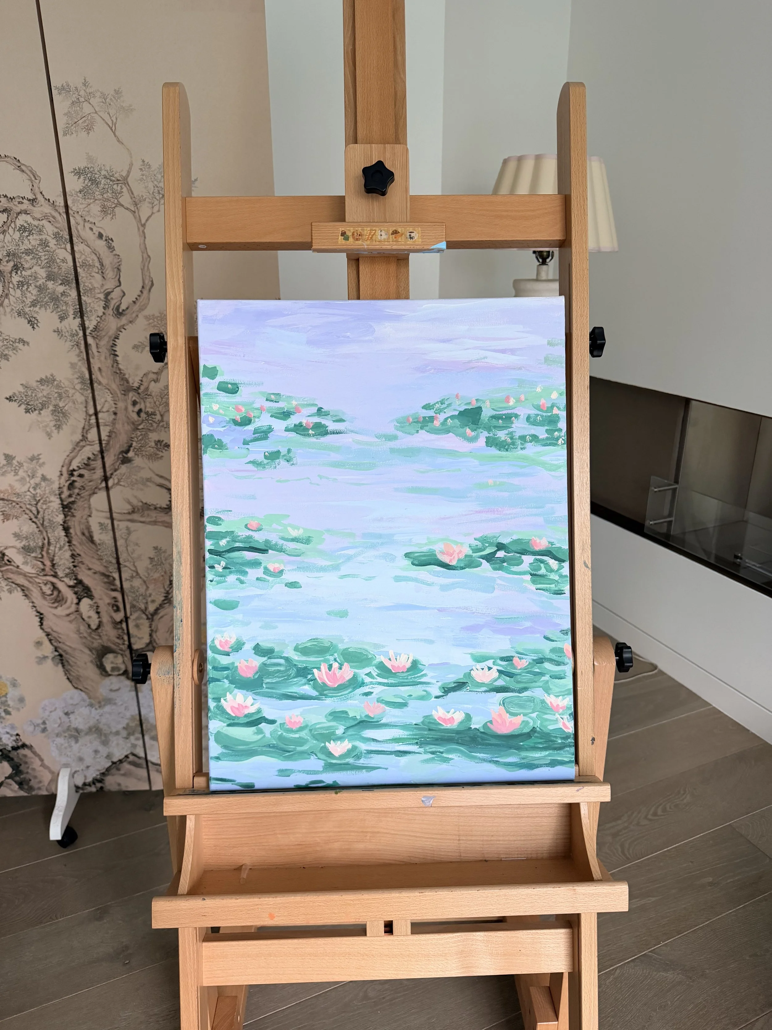 An easel displaying a painting of a water scene with green lily pads and pink water lilies.