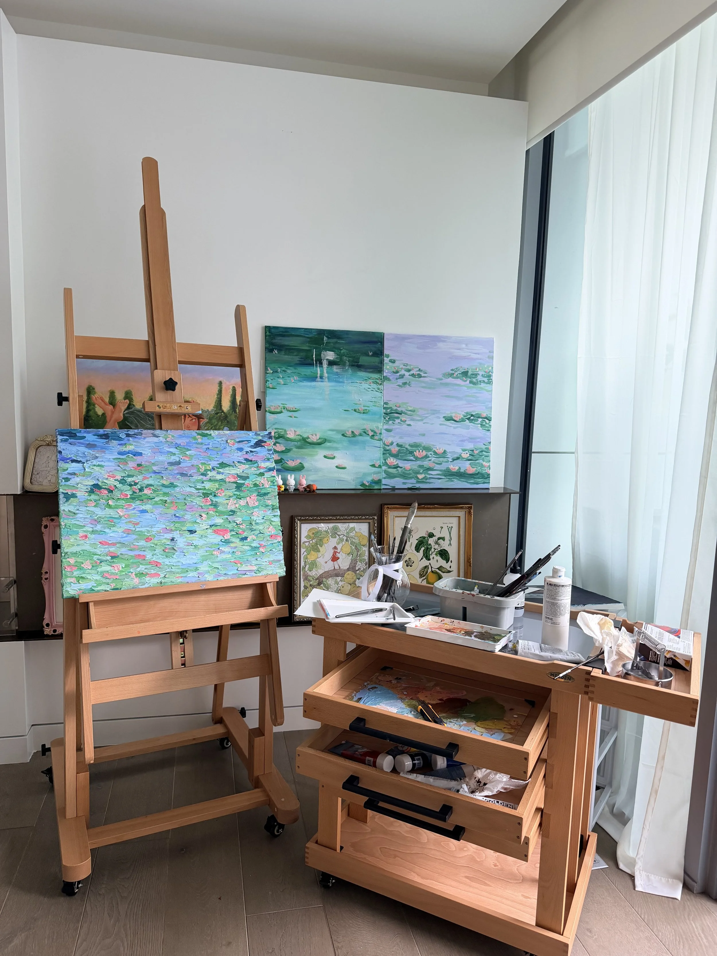 An artist's studio with paintings, easels, and art supplies. Multiple completed water lily paintings are displayed on the wall, and an unfinished painting is on an easel. The workspace includes brushes, paints, and sketches arranged on a table and wooden carts, with a large window with curtains allowing natural light.