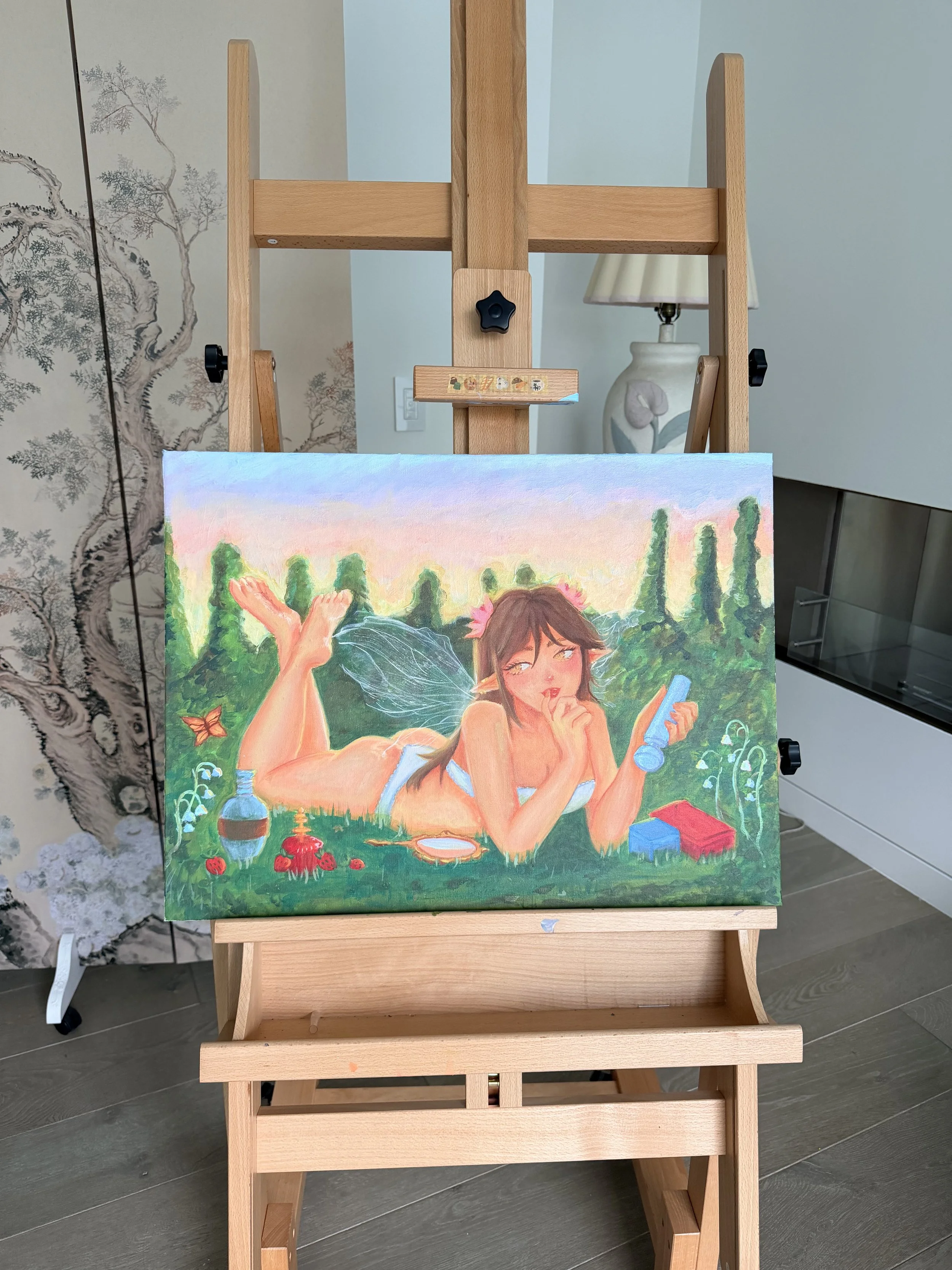 A wooden easel displaying a colorful painting of a woman lying on grass with trees and a sunset background, surrounded by art supplies and natural scenery.