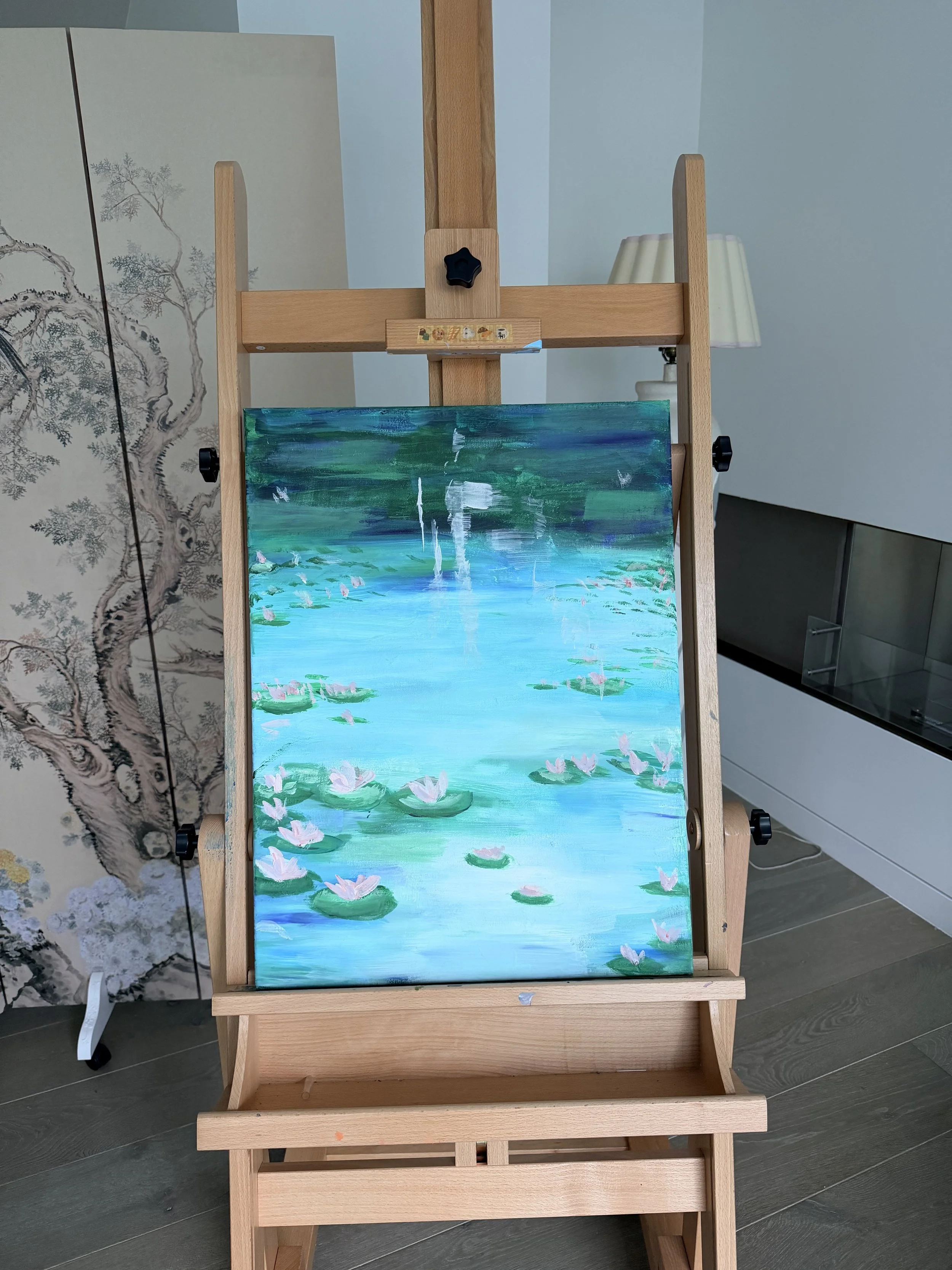 An easel holding a painting of a water lily pond scene with pink water lilies, green lily pads, and reflections on the water. In the background are a painted screen with a tree design, a ceiling light, and a white cabinet.