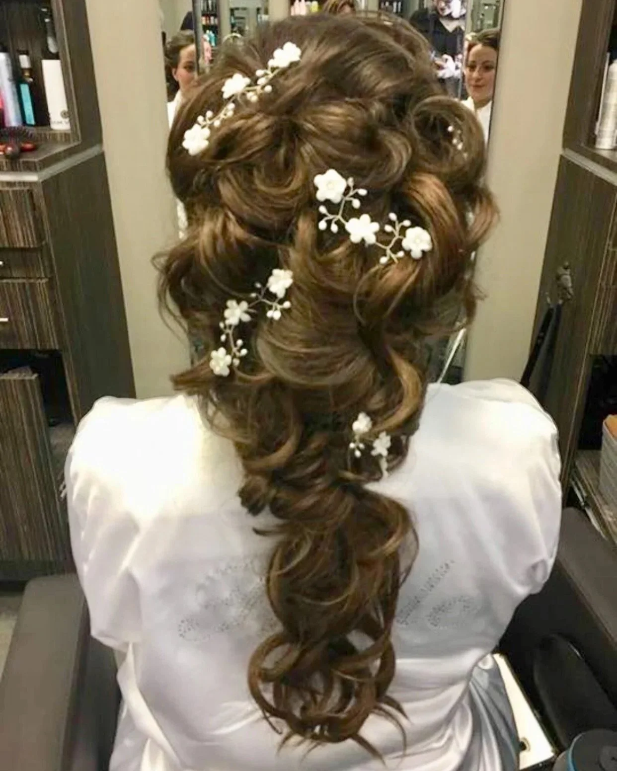 Back view of a woman with long, styled, curly hair adorned with white floral hair accessories, sitting in a salon or dressing room.