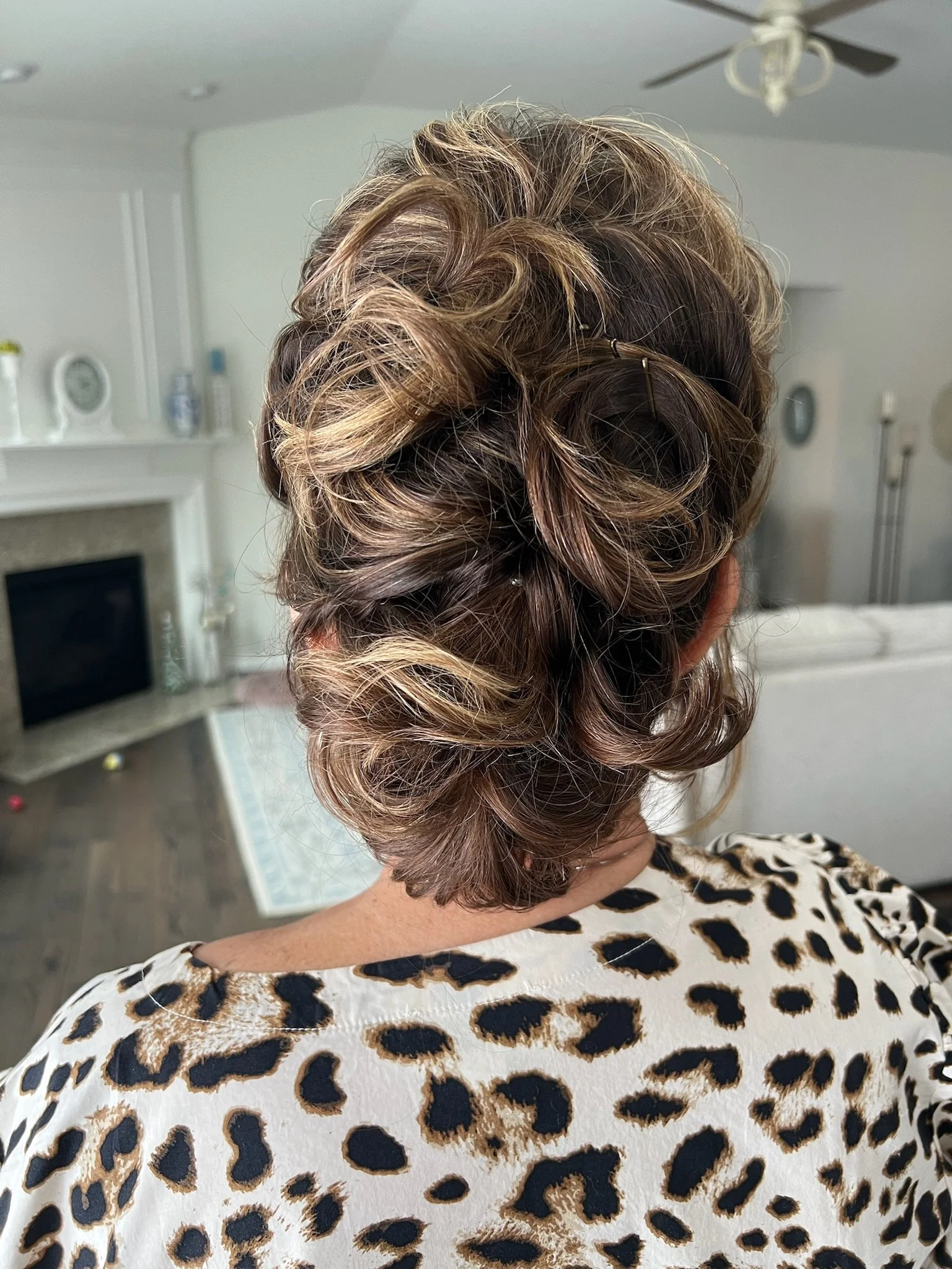 Back of a woman's head with styled, curly, blonde and light brown hair, wearing a leopard print top in a room with a ceiling fan and a fireplace.