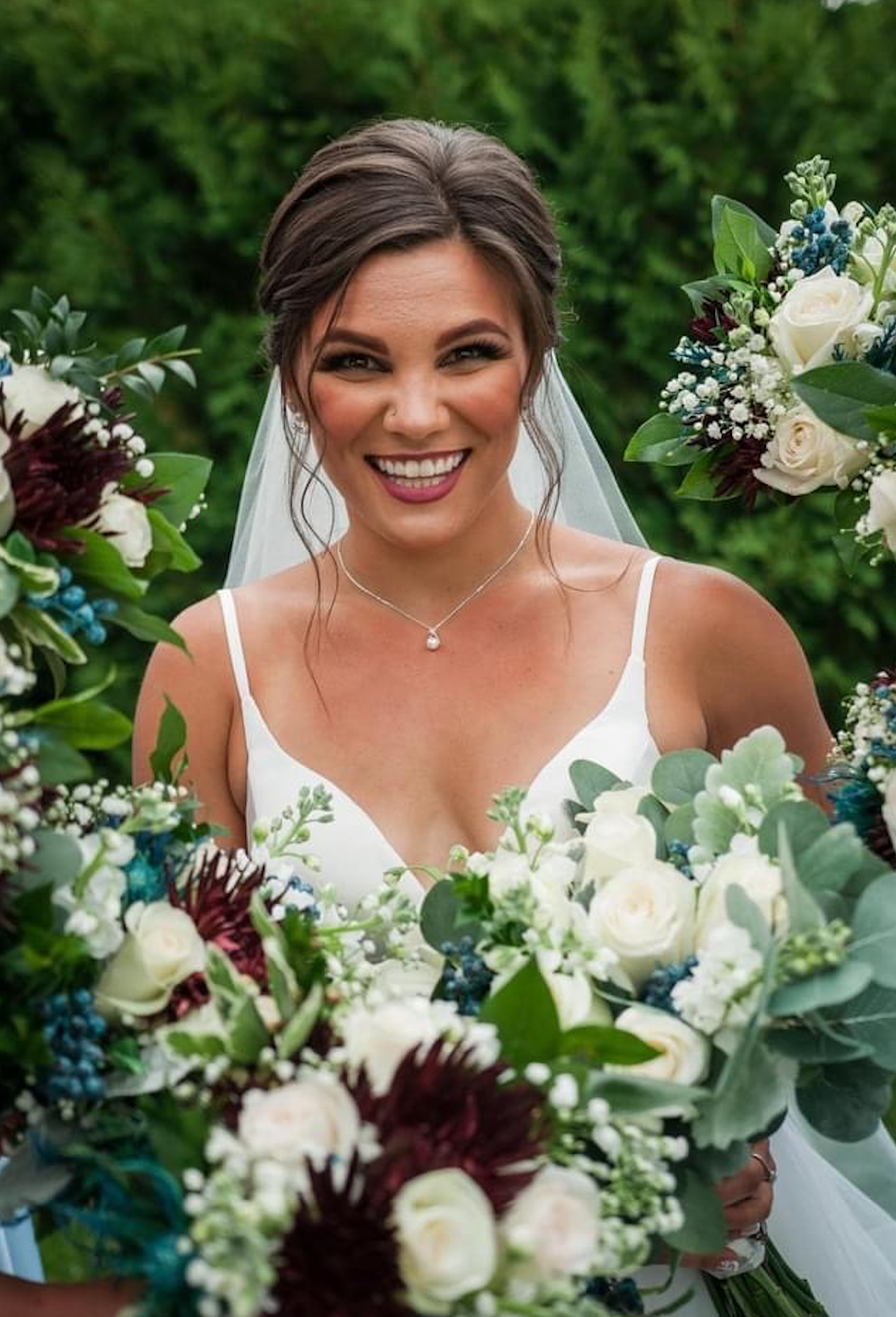 A bride with brown hair styled in an updo, smiling, wearing a white dress with thin straps, a necklace, and veil, surrounded by a large floral bouquet with white, purple, and green flowers.