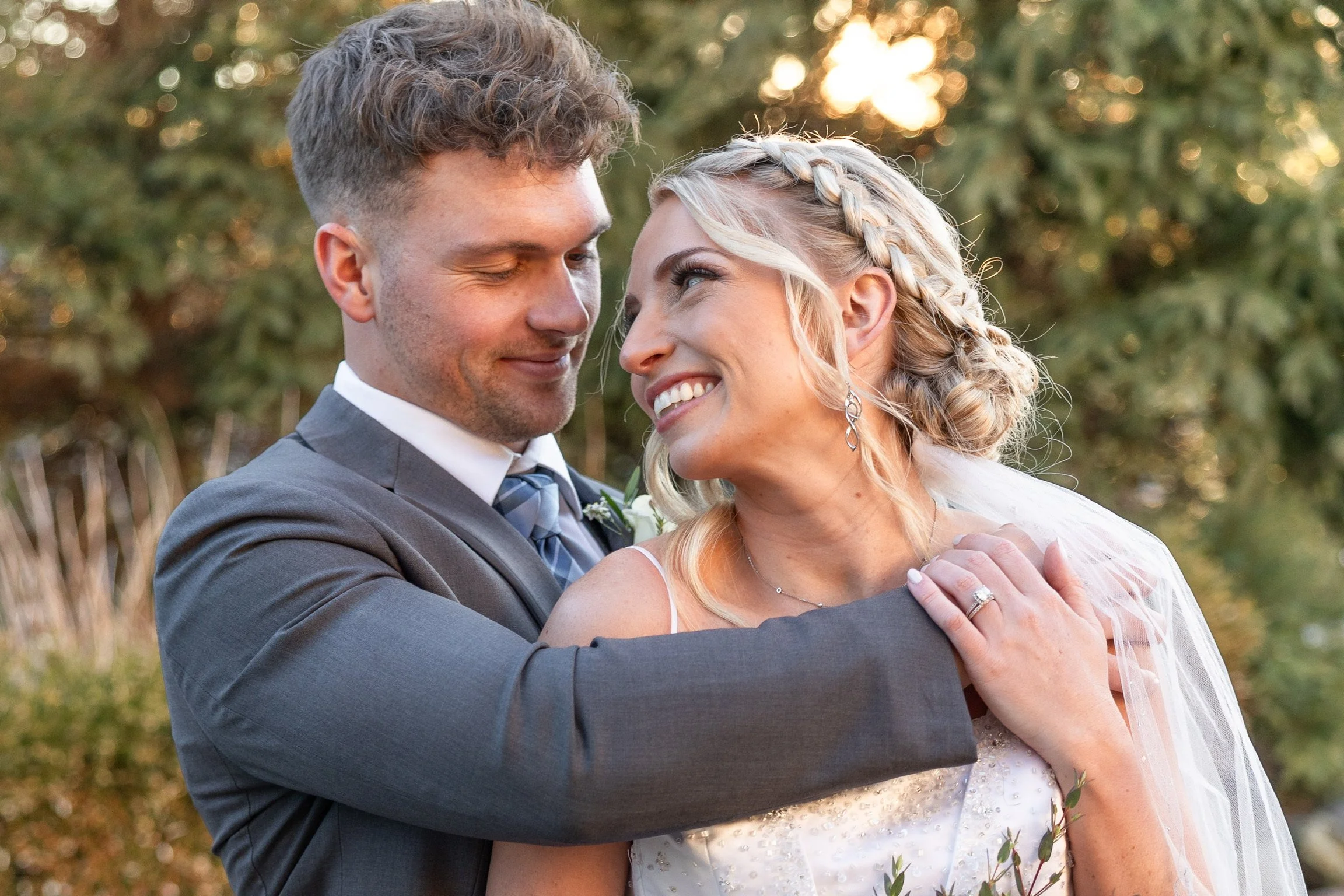 A newlywed couple is embracing outdoors during sunset, with the groom in a gray suit and the bride in a white wedding dress with lace details and braided blonde hair.